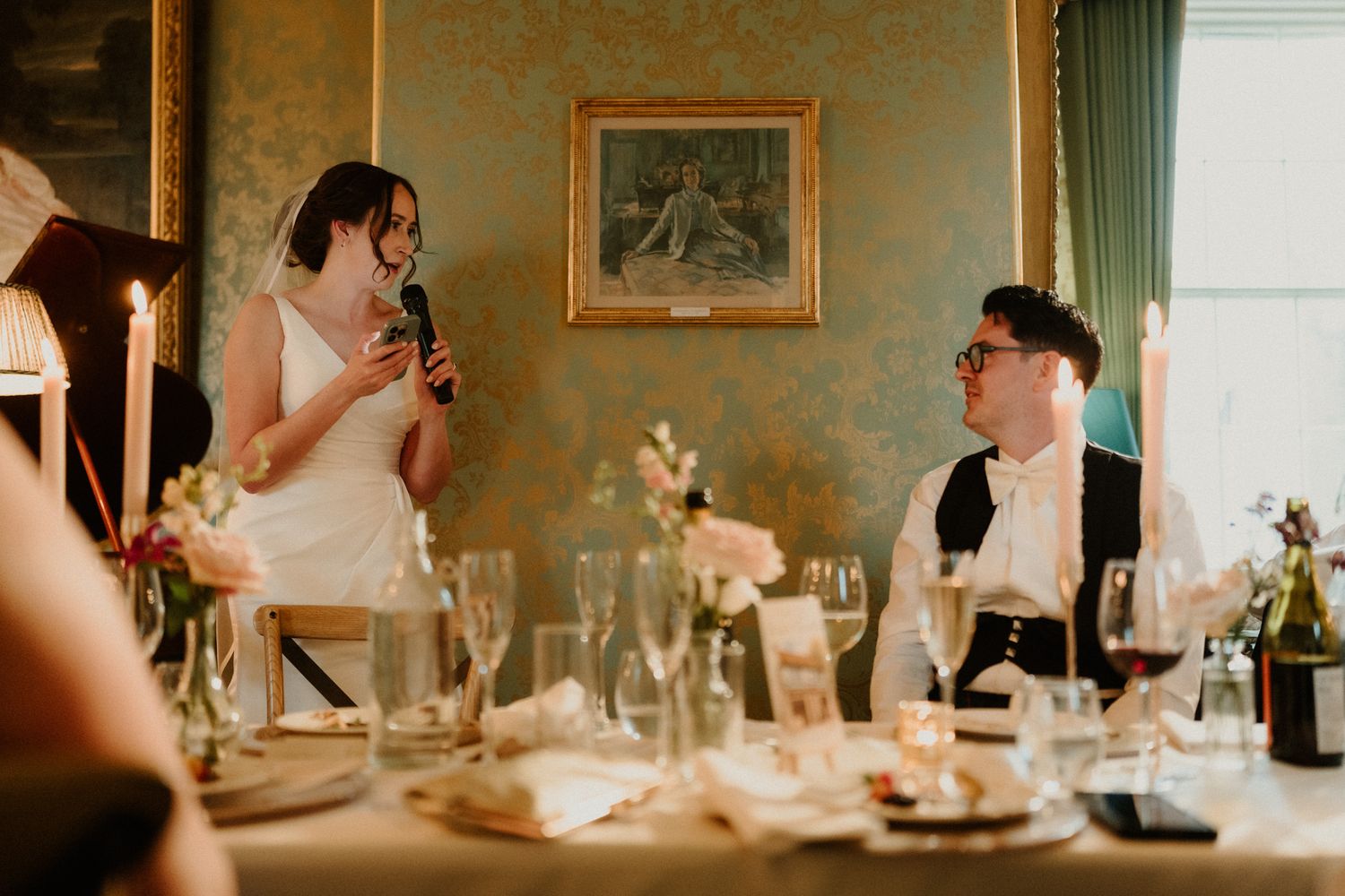 Bride in a white dress speaks into a microphone at a wedding reception table with candles, flowers and glasses as a smiling groom listens.