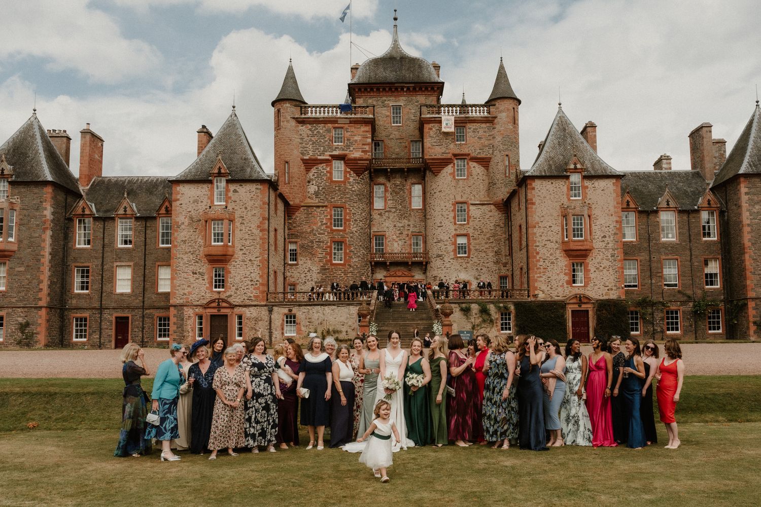 Wedding guests in colorful formal dresses pose for a group photo on a lawn in front of a historic red sandstone Scottish castle.