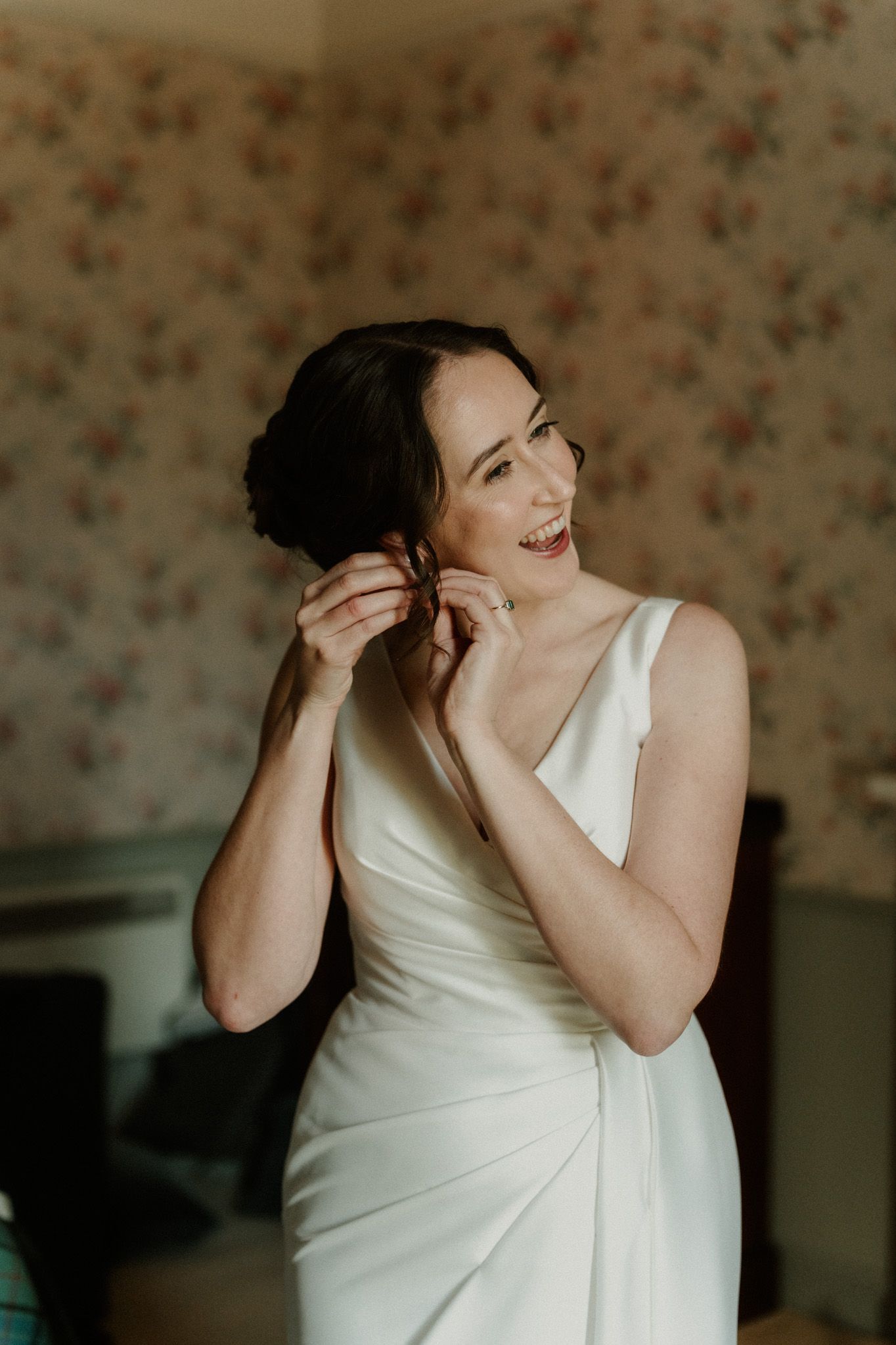 Smiling bride in an elegant white satin gown putting on an earring while getting ready in a room with floral vintage wallpaper.