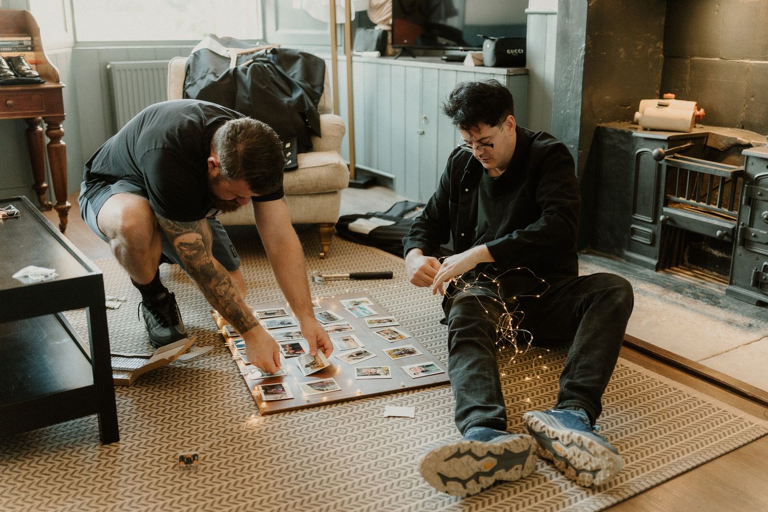 Two men sit on a woven rug in a bright room, arranging and examining printed photos spread out on a wooden board.