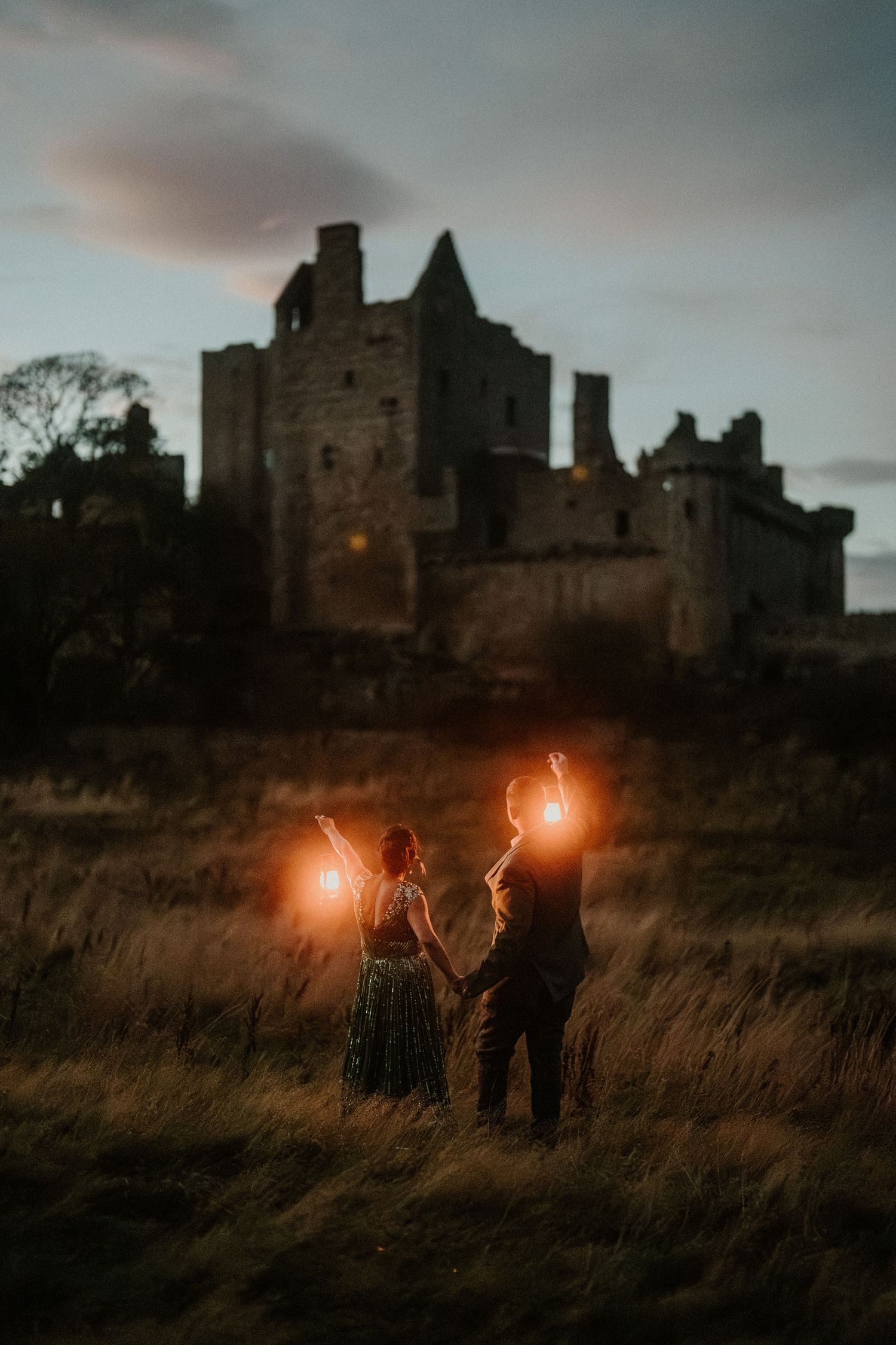Couple holding glowing lanterns in a field at dusk before a historic castle ruin at twilight.