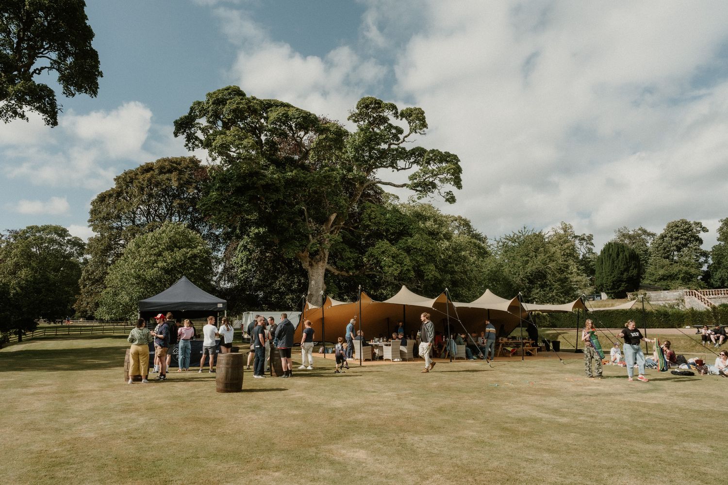 Guests mingle around tents and a dark canopy on a sunny lawn beneath large leafy trees at an outdoor garden party or wedding reception.