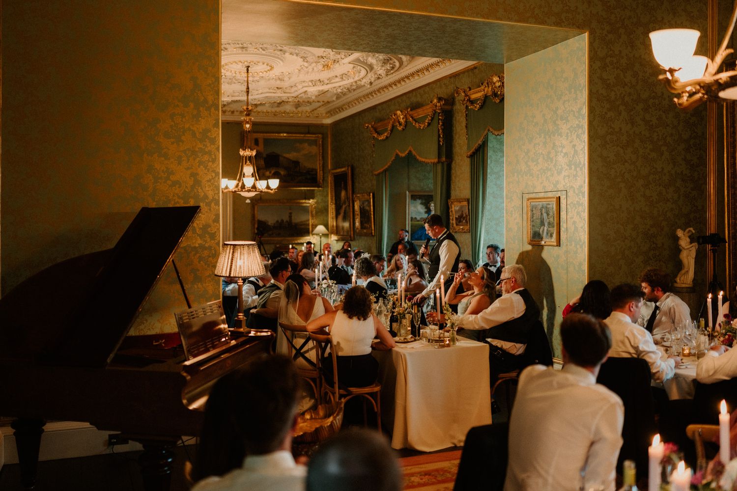 Guests dine by candlelight at elegant tables in a grand ballroom with chandeliers, ornate ceilings, green walls and a black grand piano.