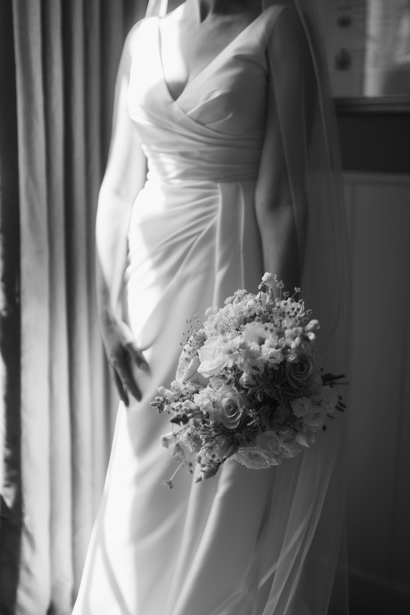 Black and white photo of a bride in a wrap-style satin wedding gown holding a bouquet of roses beside a curtained window.
