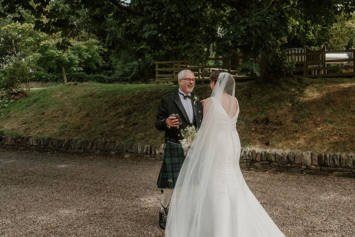 Smiling groom in a kilt and dark jacket greets his veiled bride in a long white wedding gown on a gravel path outdoors.