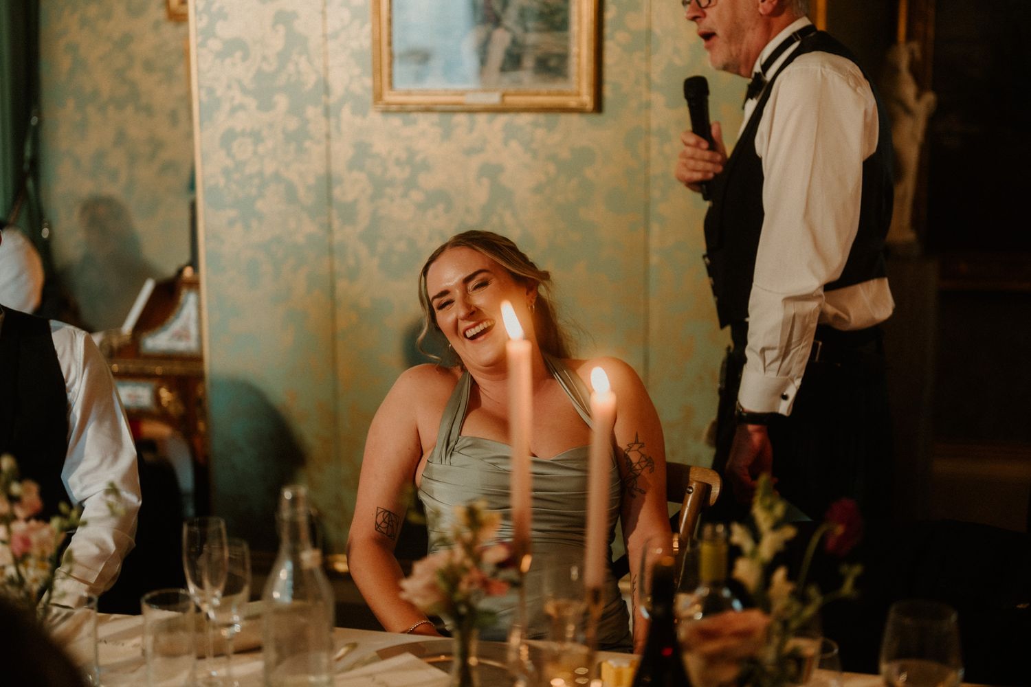 A woman laughs joyfully at a candlelit dinner table as a man in a vest speaks into a microphone in an elegant room with floral wallpaper.