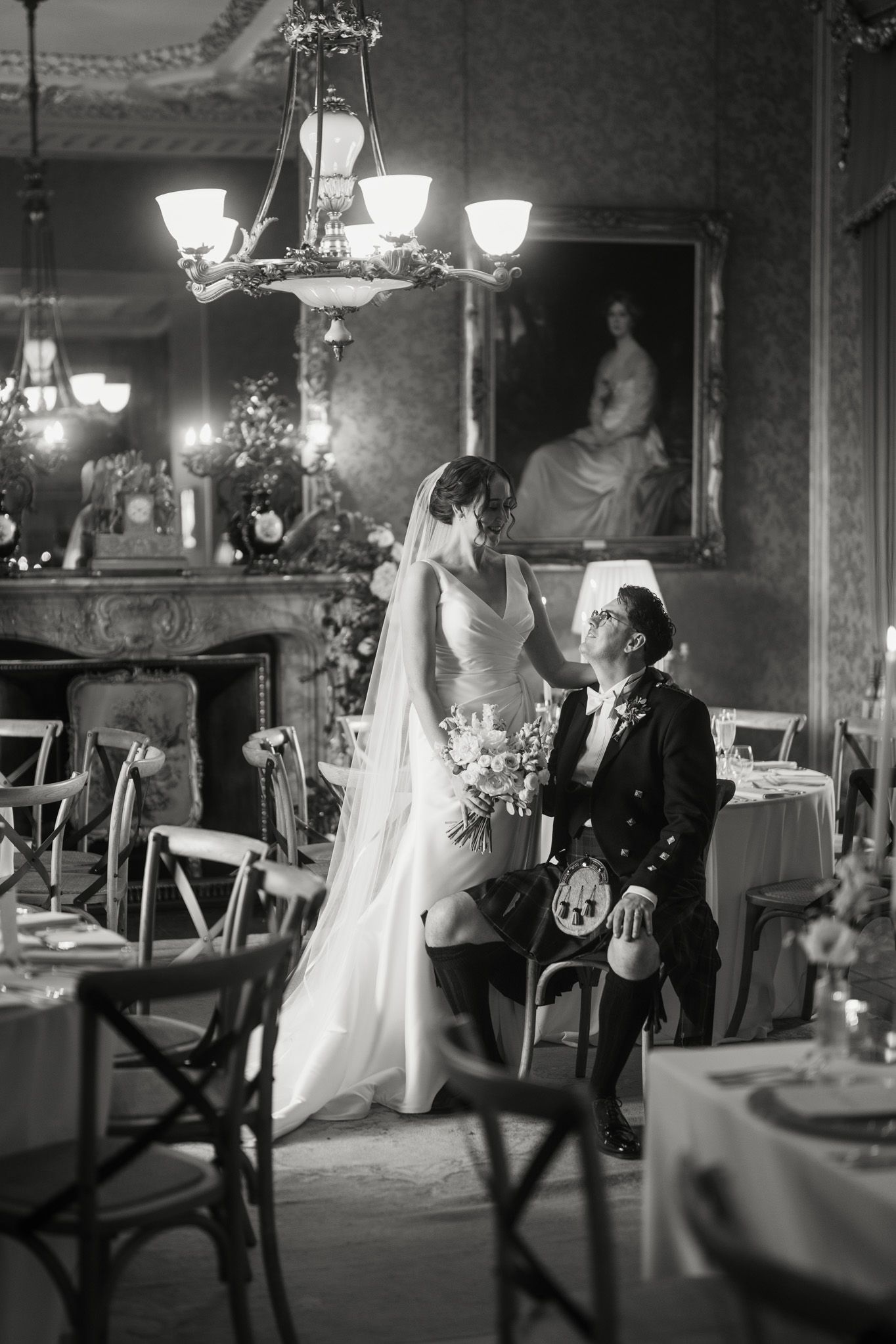 Black and white wedding photo of a bride in a flowing veil standing beside a seated groom in a kilt in an elegant reception room.