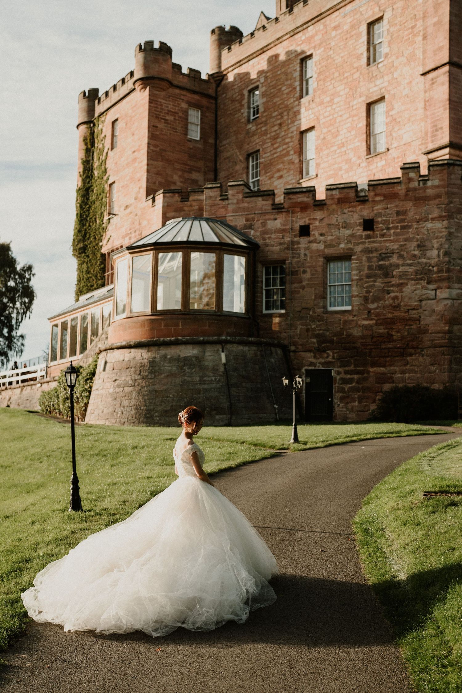 A dramatic wedding dress billows on the path leading to a historic castle.