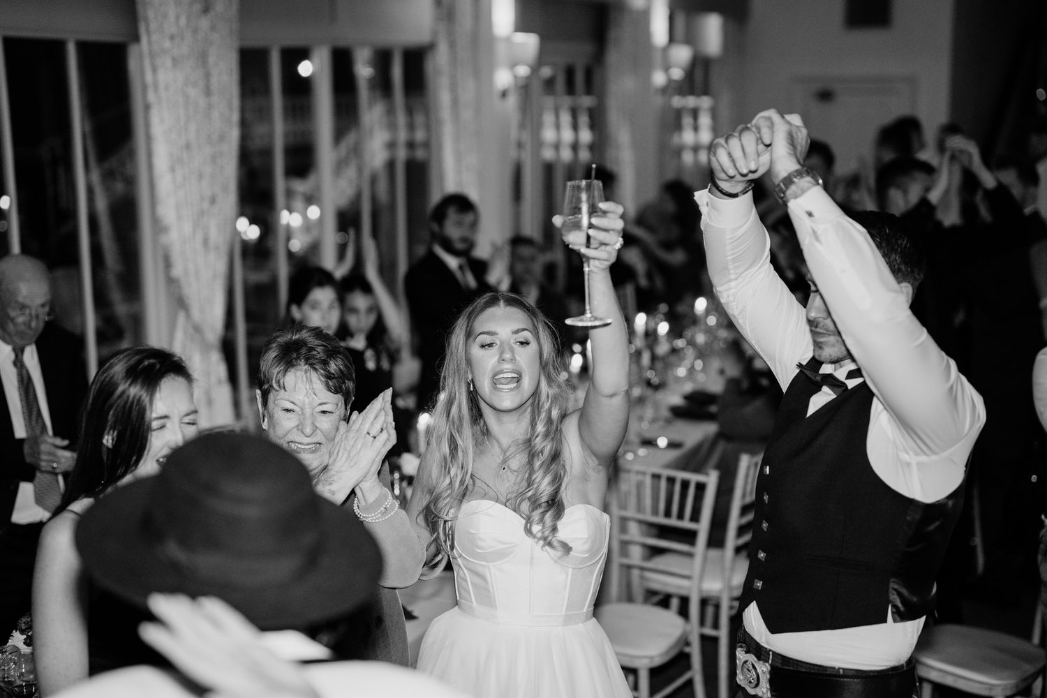 Black and white photo of wedding guests celebrating at a reception with hands raised.