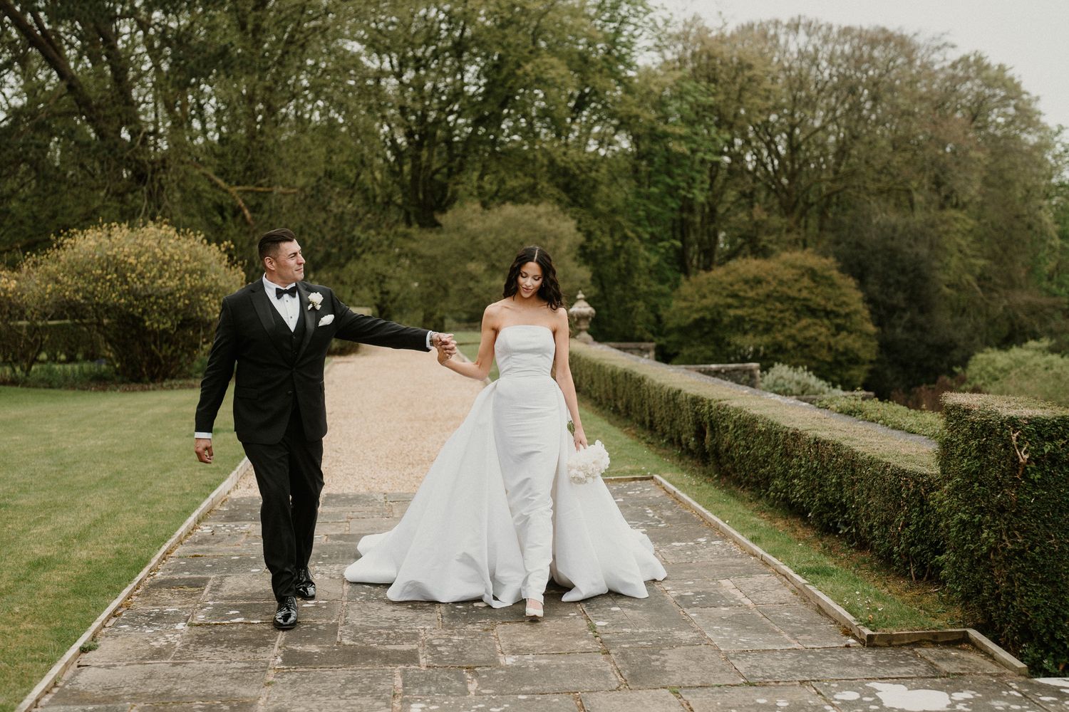 A joyful wedding couple strolls down a garden path surrounded by spring foliage.