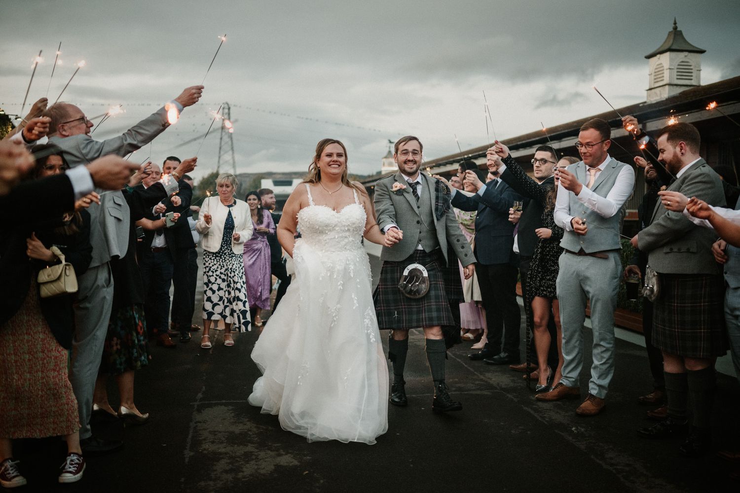 Wedding guests create a celebratory exit path with confetti against a cloudy sky.