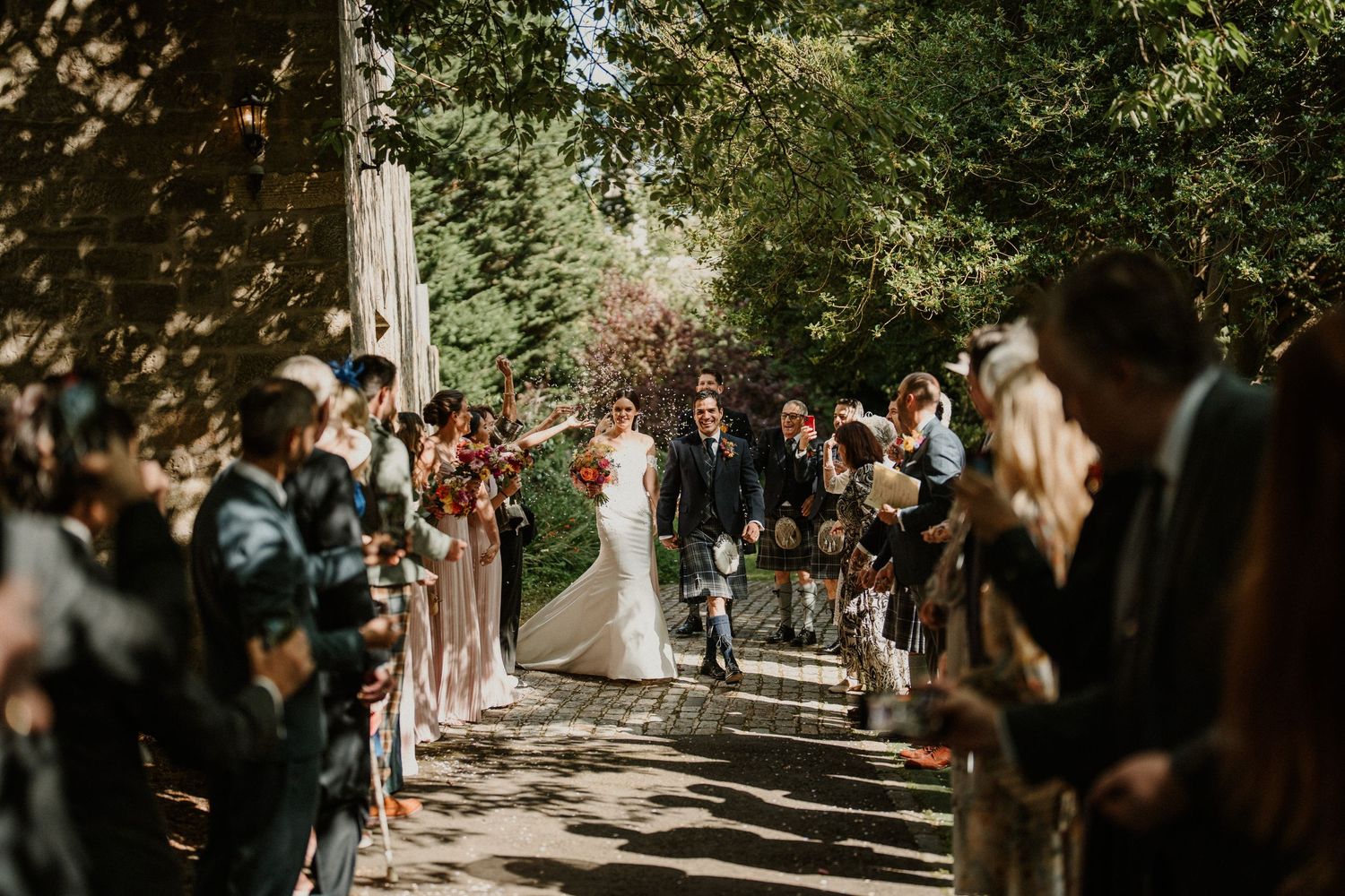 Wedding guests gathered outdoors under tree-lined path for ceremony.