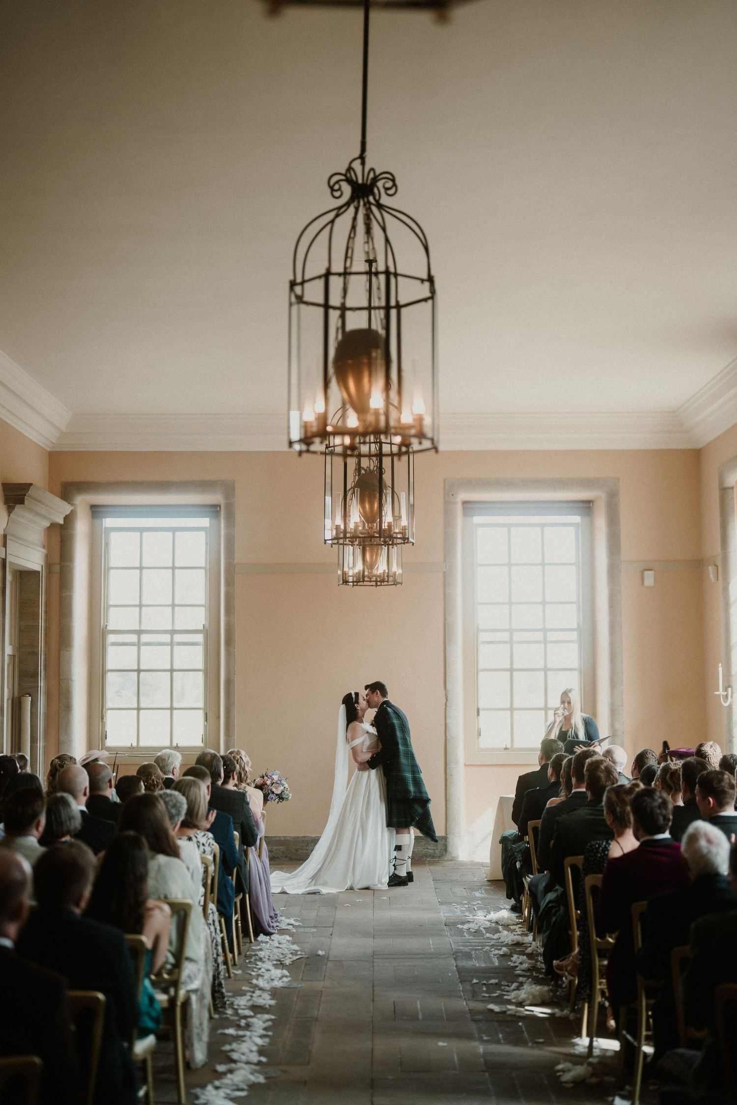 Wedding ceremony in progress in a bright room with hanging chandeliers and tall windows.