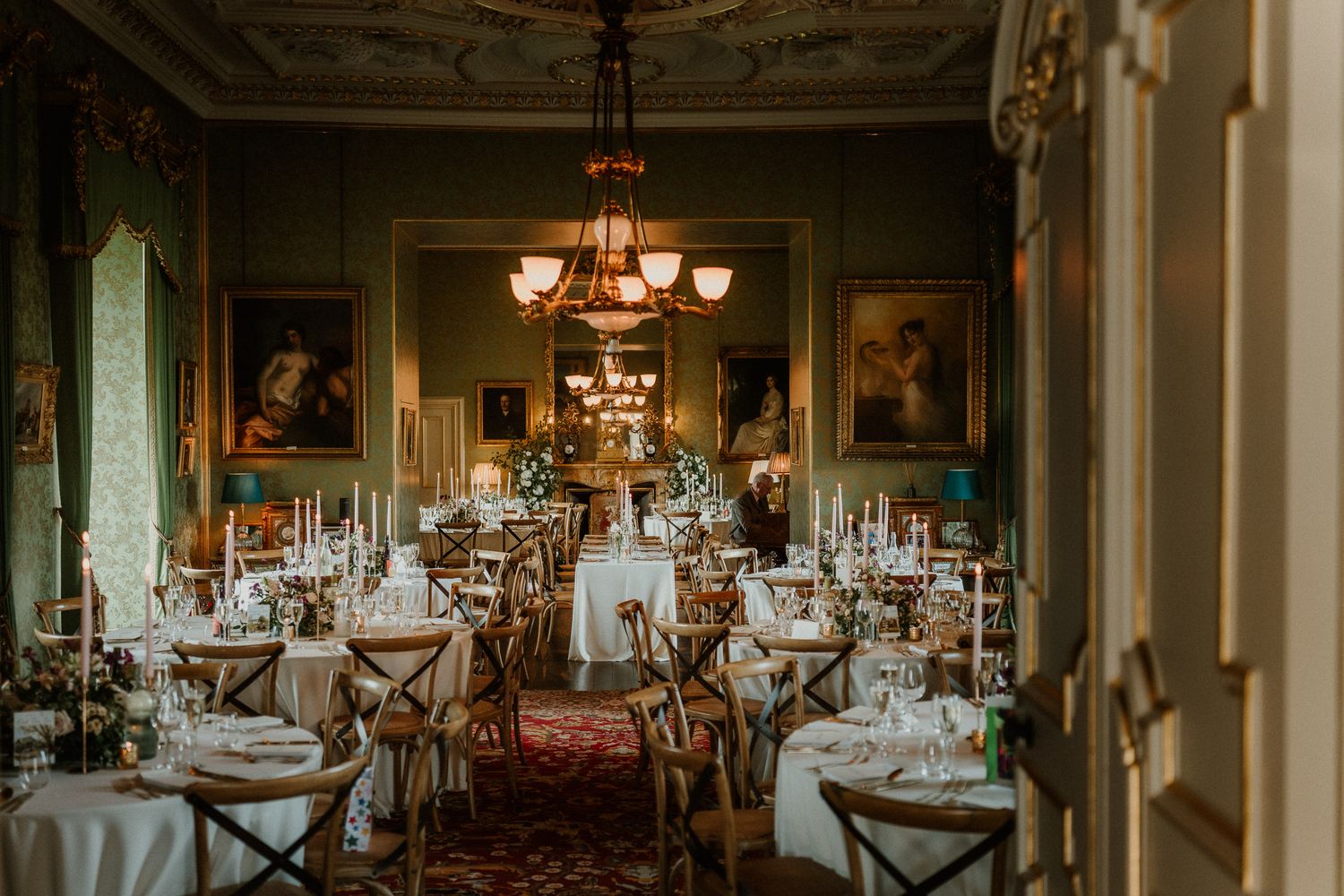 Elegant wedding reception in a grand room with chandeliers, white tablecloths, cross-back chairs, and ornate paintings on green walls.