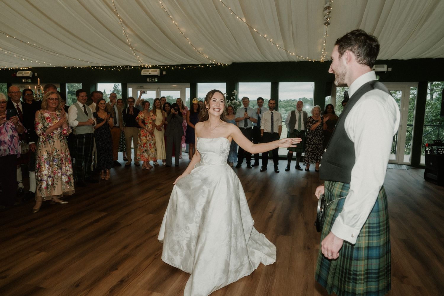 Bride in silver gown and groom in tartan kilt share first dance at elegant wedding reception.