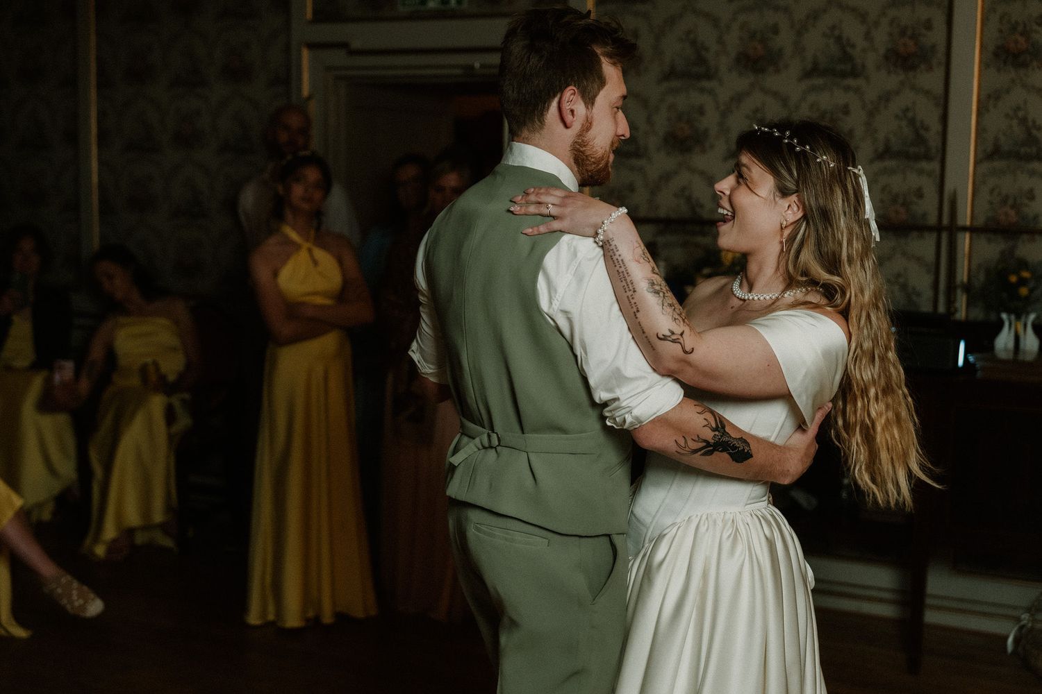 Bride and groom share their first dance at wedding reception, guests in yellow dresses watch in background.
