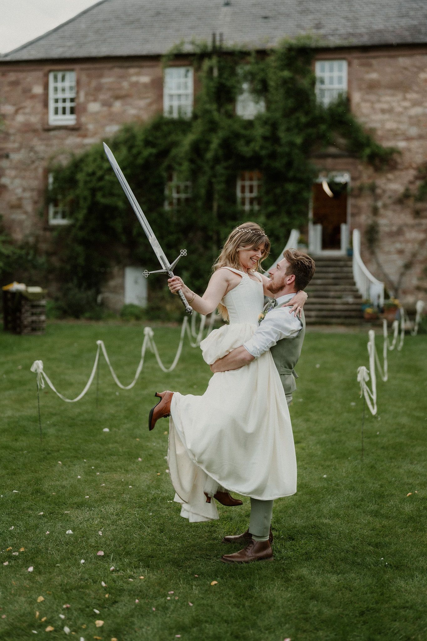 Groom lifts bride holding a sword outside a stunning ivy-covered stone wedding venue.