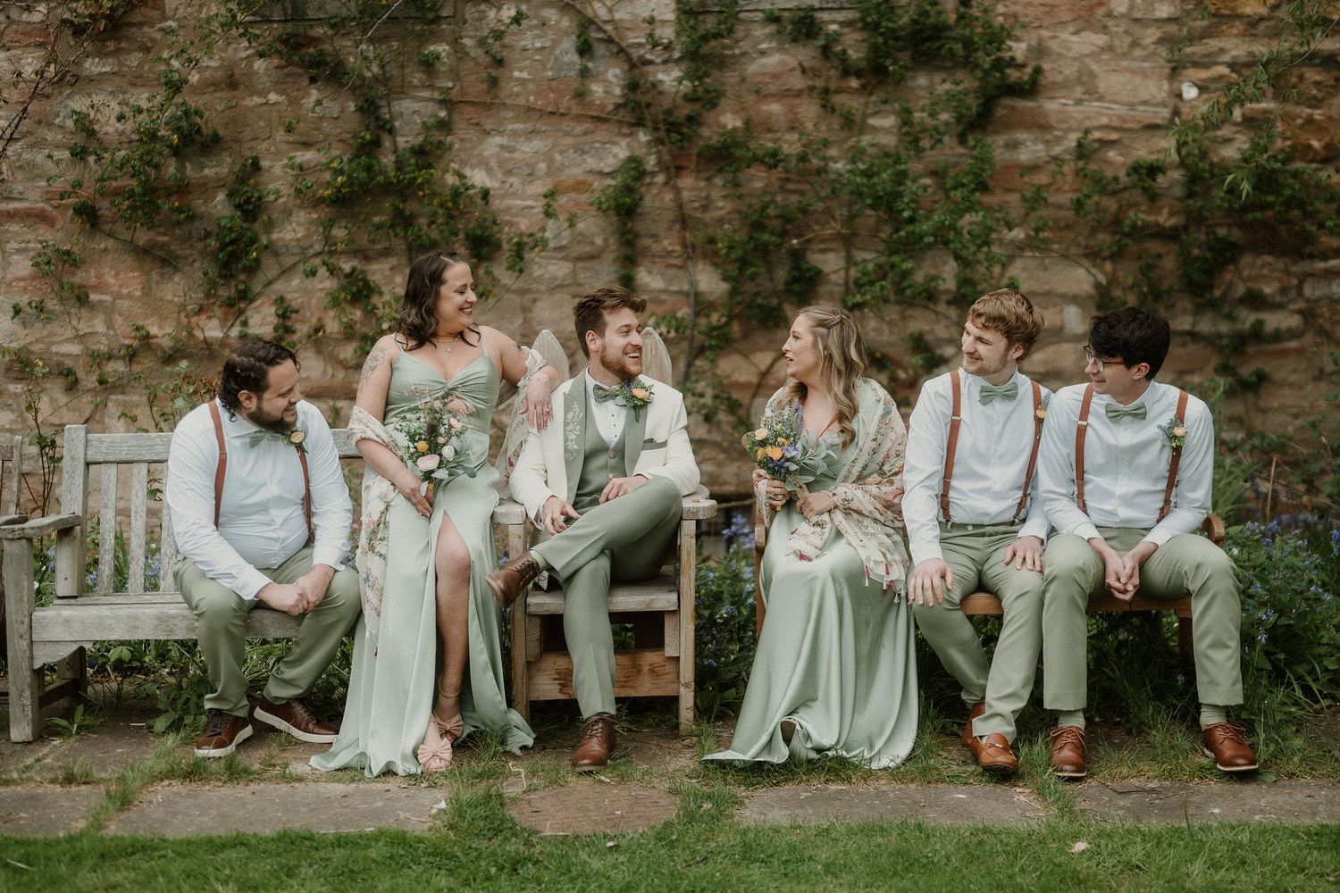 Wedding party in sage green attire laughing together against a rustic stone wall with a wooden bench outdoors.
