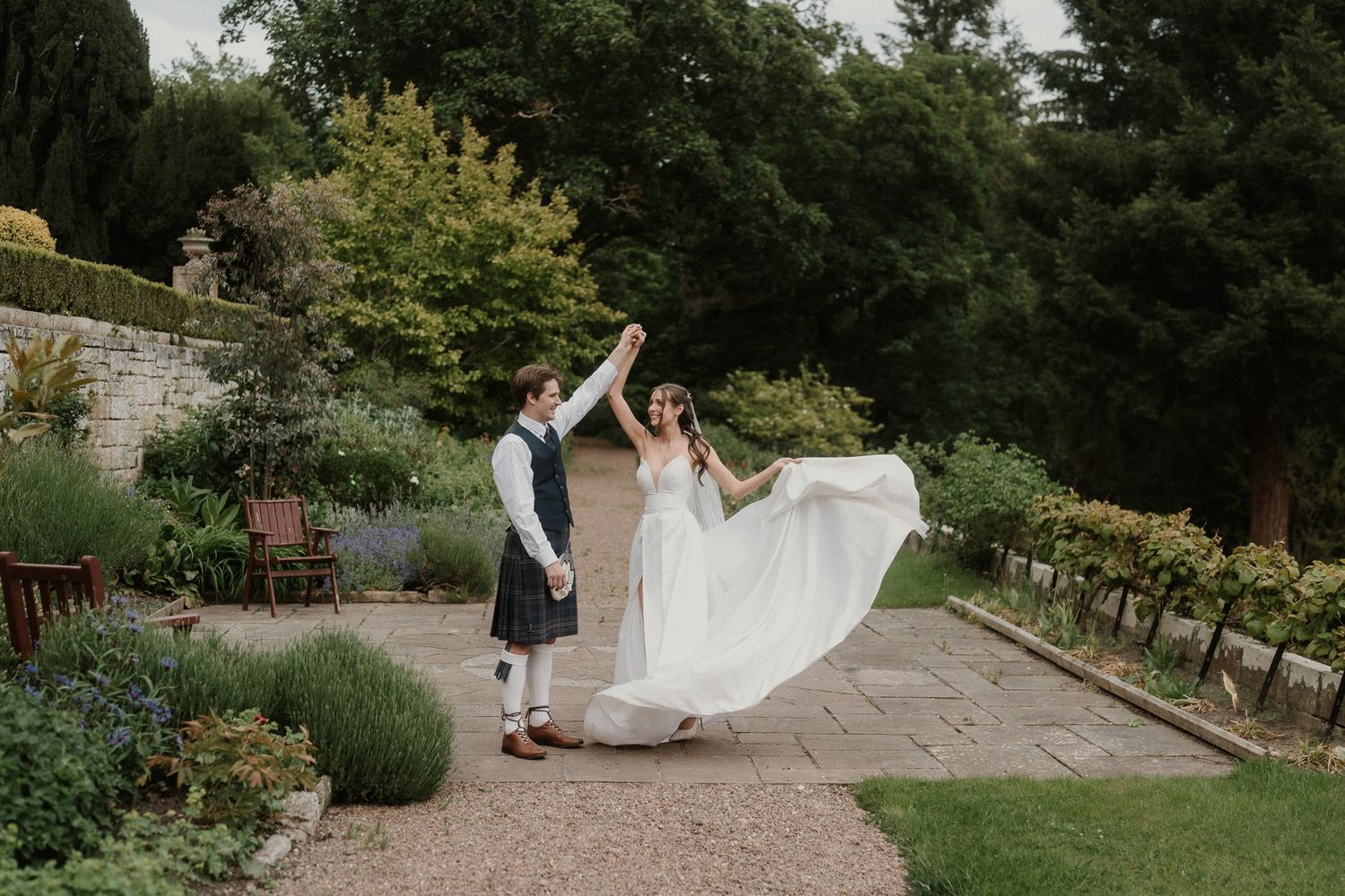 Bride twirling in white gown with groom in kilt on garden path at outdoor wedding venue.
