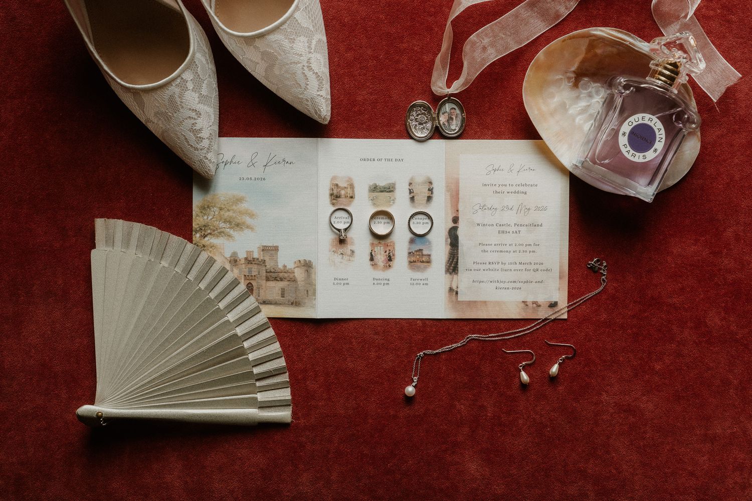 Elegant wedding flat lay with lace bridal shoes, rings, invitations, perfume, fan, and pearl earrings on red velvet.