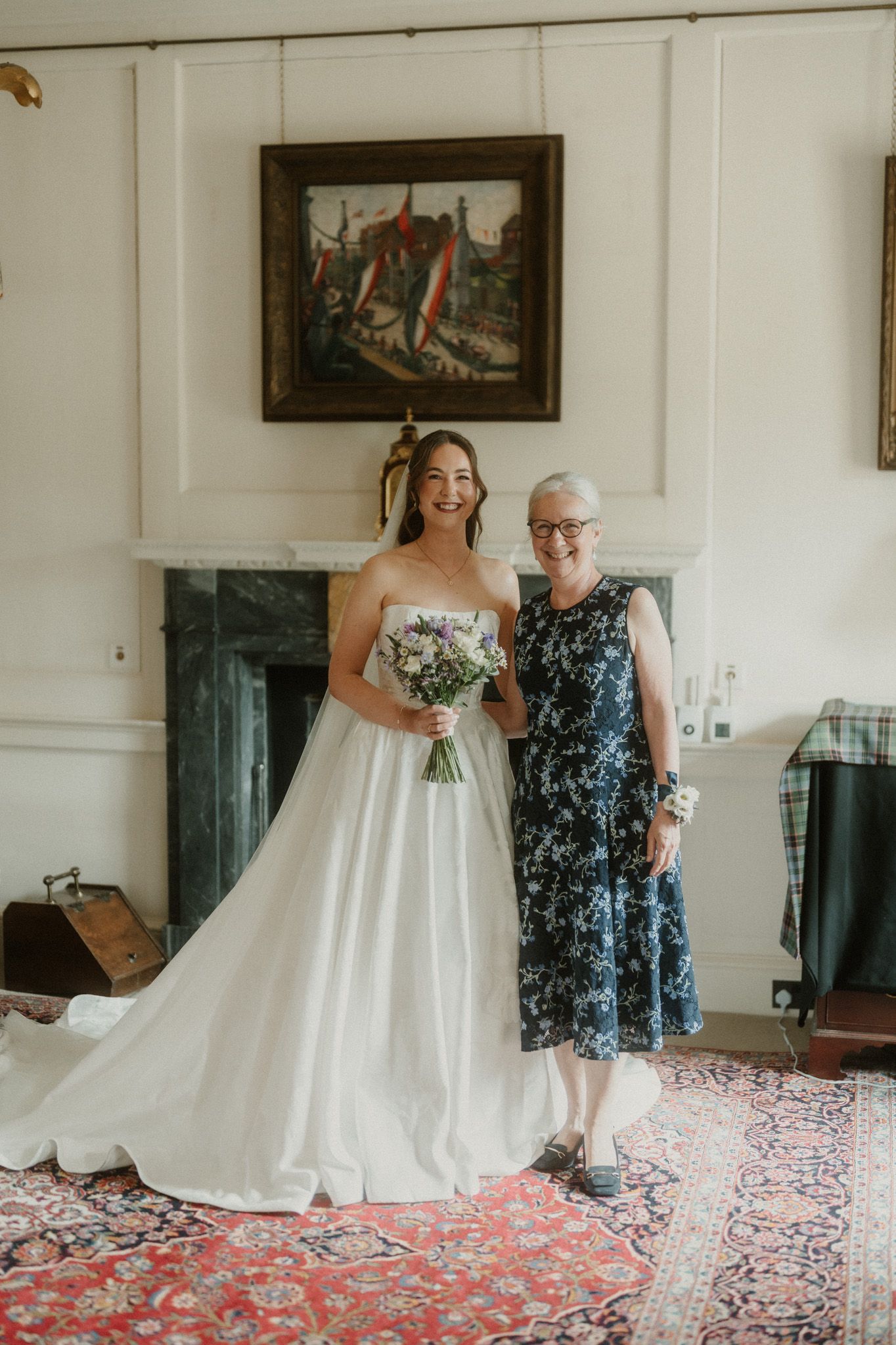 Bride in white gown holding bouquet poses with older woman in blue floral dress before a fireplace.