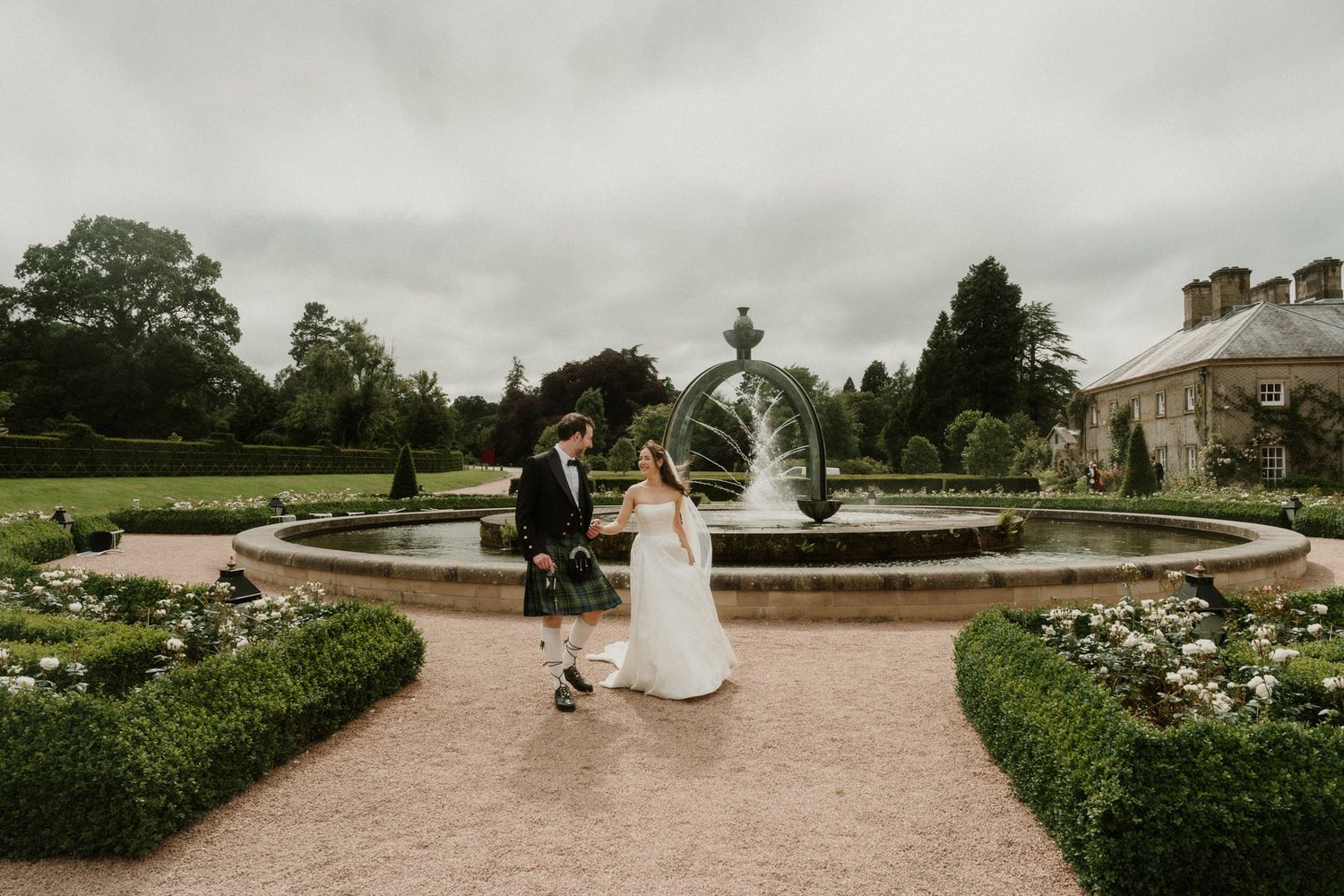 Bride and groom walk hand in hand by a fountain in a formal garden at a Scottish wedding venue.