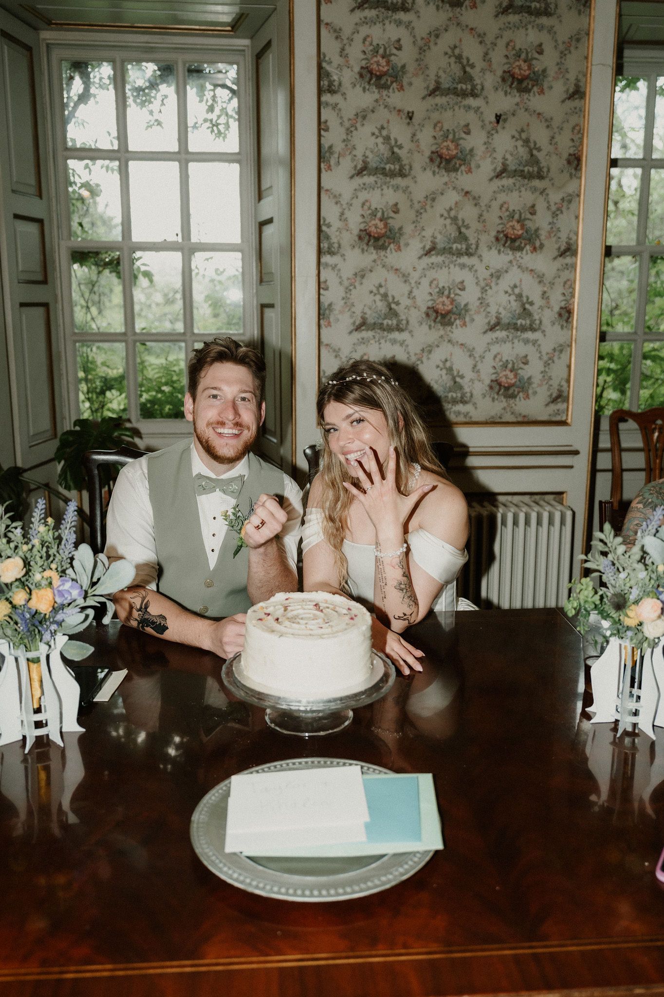 Smiling newlyweds sit at elegant table with white wedding cake, bride shows ring, floral wallpaper backdrop.