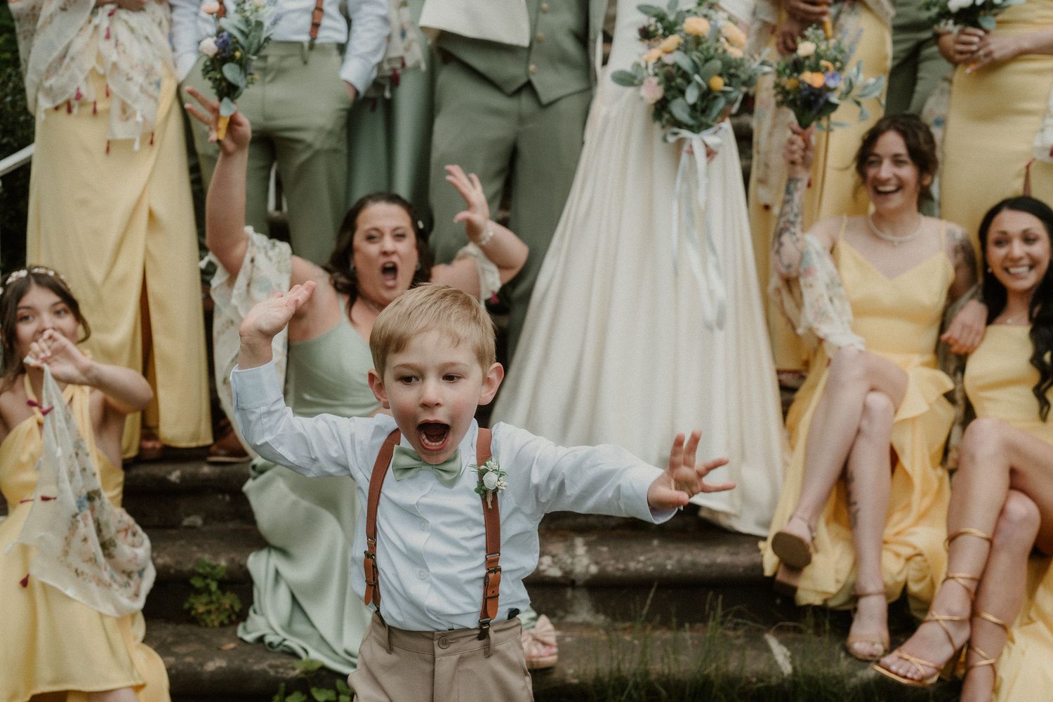 Excited young ring bearer with suspenders celebrates joyfully at a wedding with bridesmaids in yellow and green.