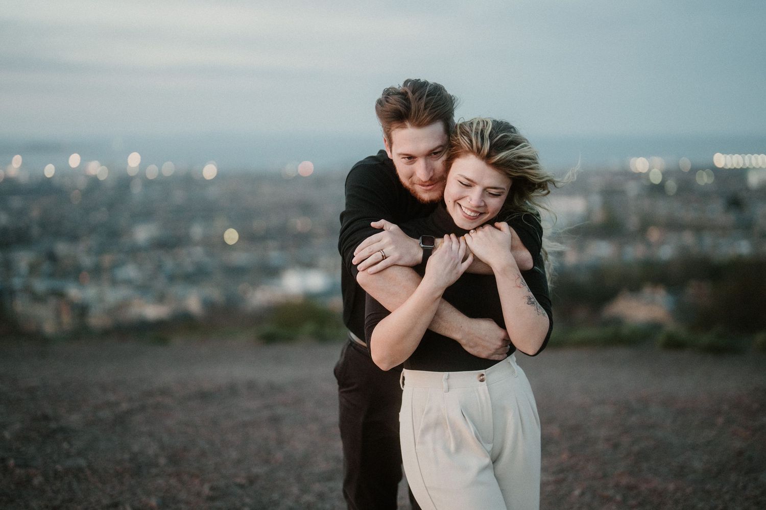 Couple embracing romantically on a hilltop at dusk with a city skyline in the background.