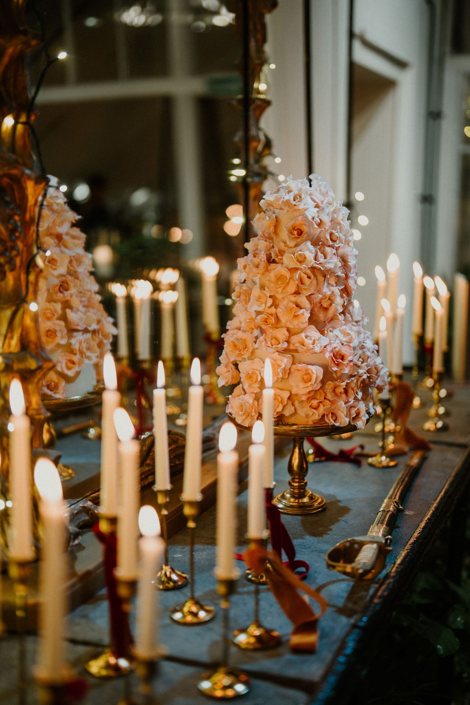 Elegant wedding cake covered in blush roses surrounded by lit white taper candles in gold holders on a decorated table.