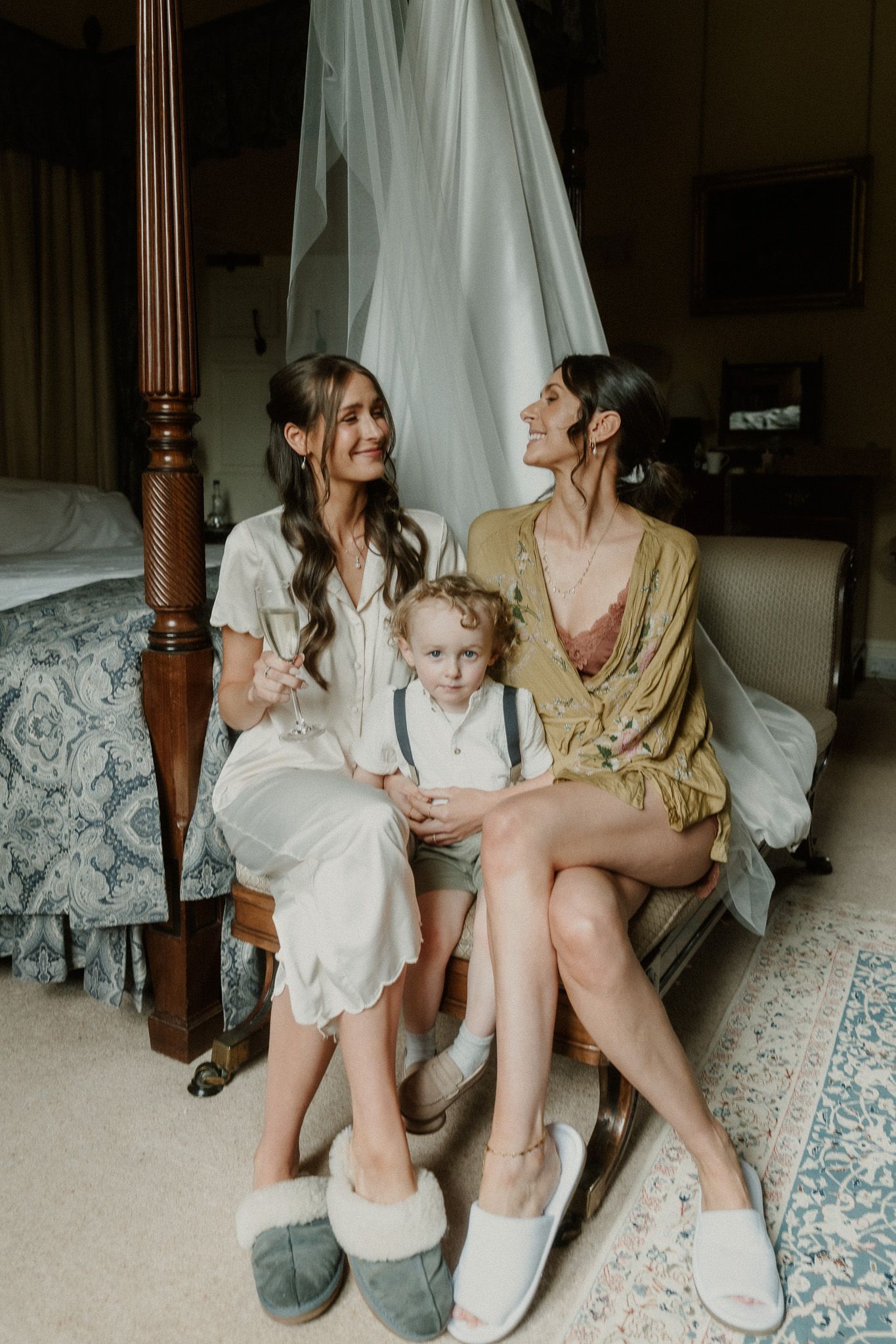 Two bridesmaids in robes sit with a young ring bearer on a bed, smiling, with a wedding dress hanging behind them.
