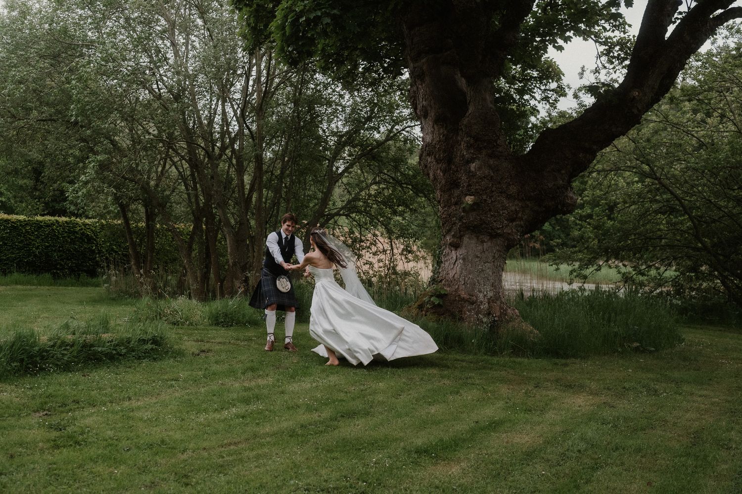 Bride and groom dancing outdoors near a large tree on lush green grass during their wedding celebration.
