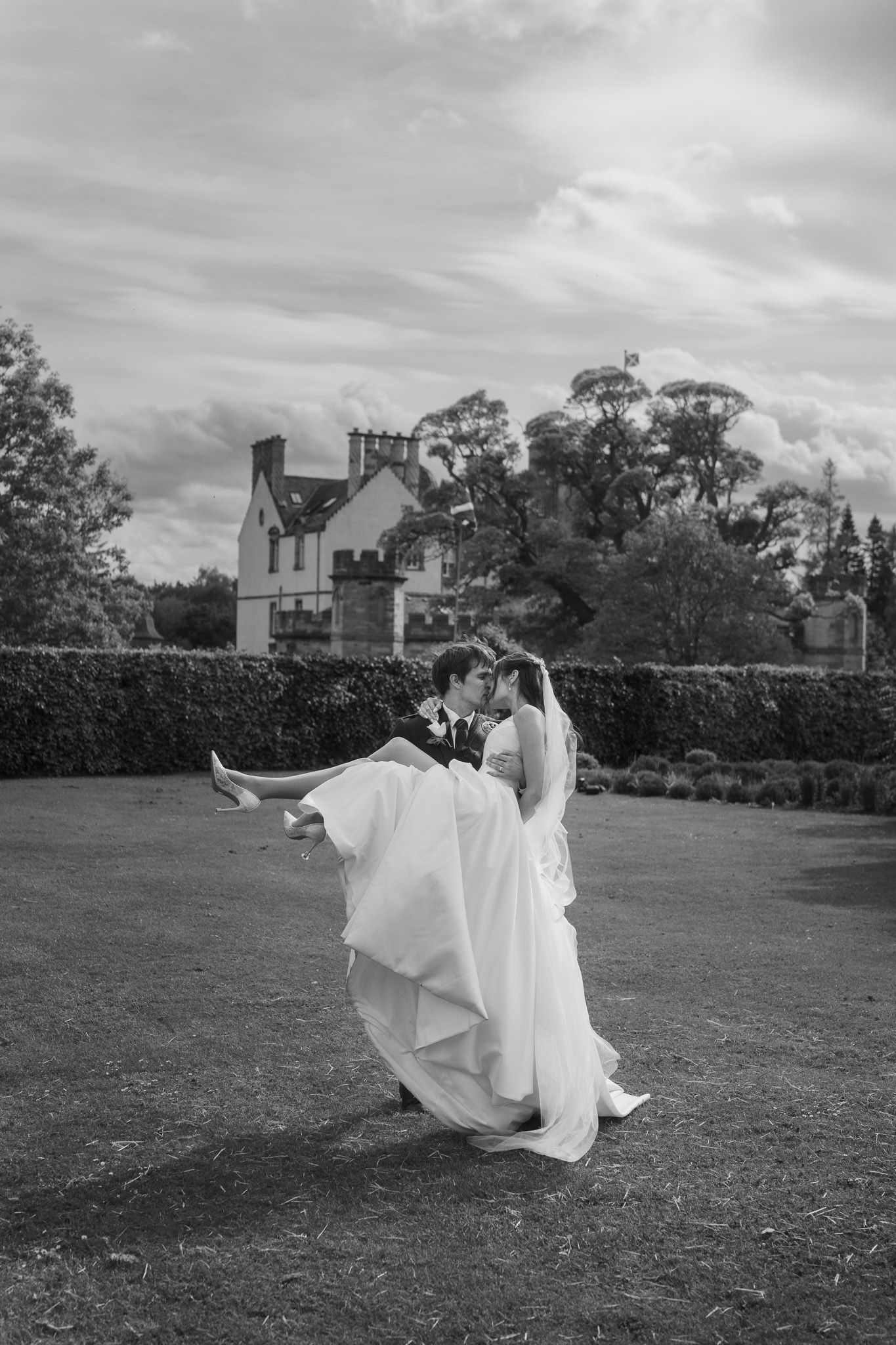 Groom carrying bride in white gown outside a grand estate, black and white wedding photo.