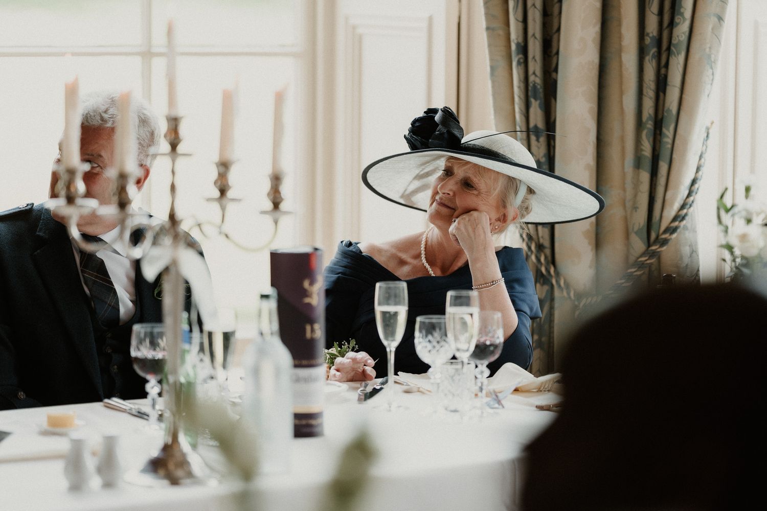 Elegant elderly woman in wide-brim hat smiling at a formal wedding reception dinner table with champagne glasses.
