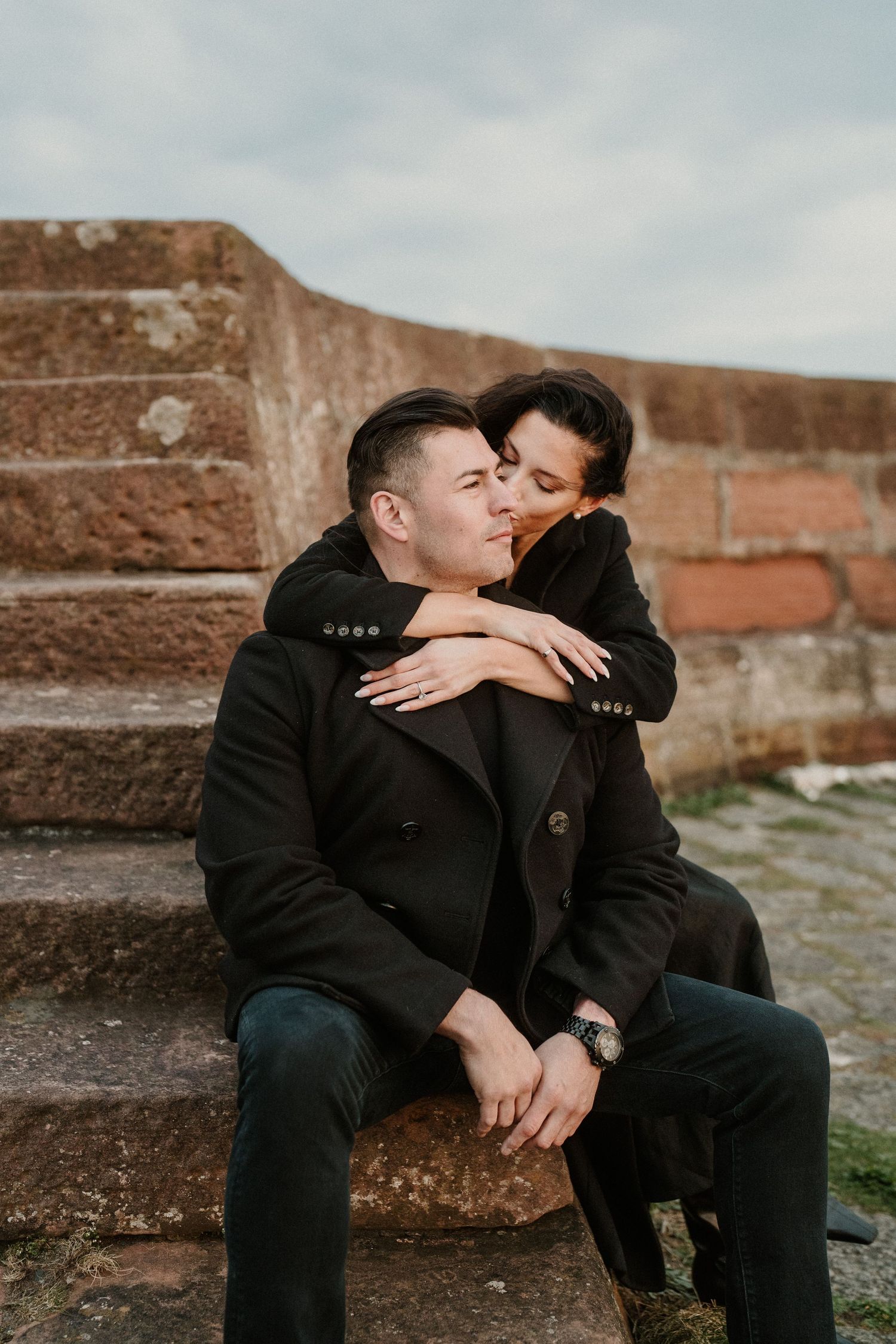 Romantic couple embracing on stone steps against an overcast sky.
