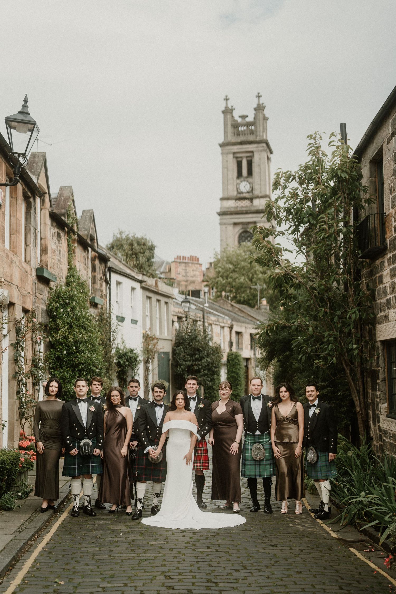 Wedding group photo on charming cobblestone street with church spire and historic buildings in background.