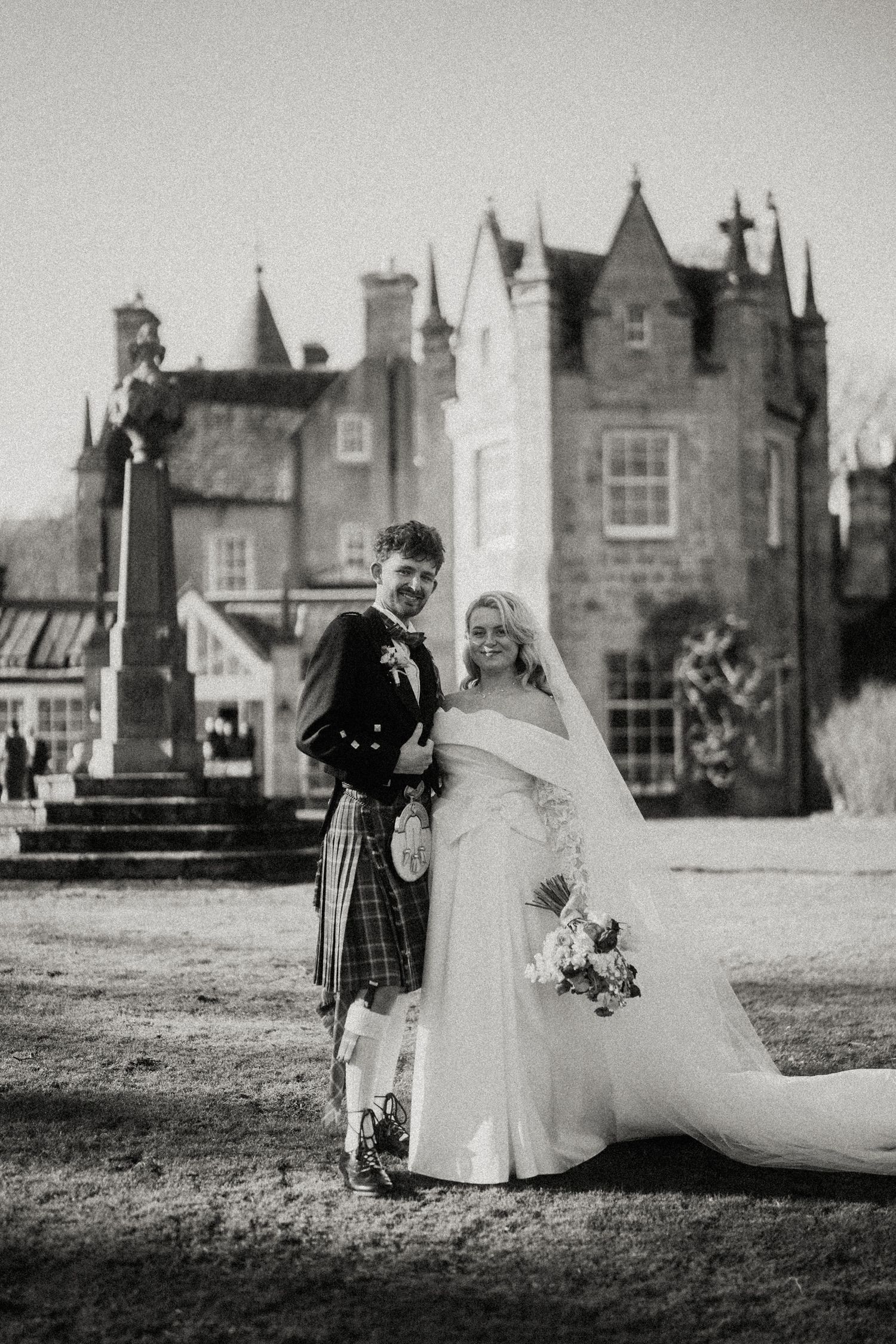 Black and white portrait of newlyweds in Scottish wedding attire standing before a historic castle.