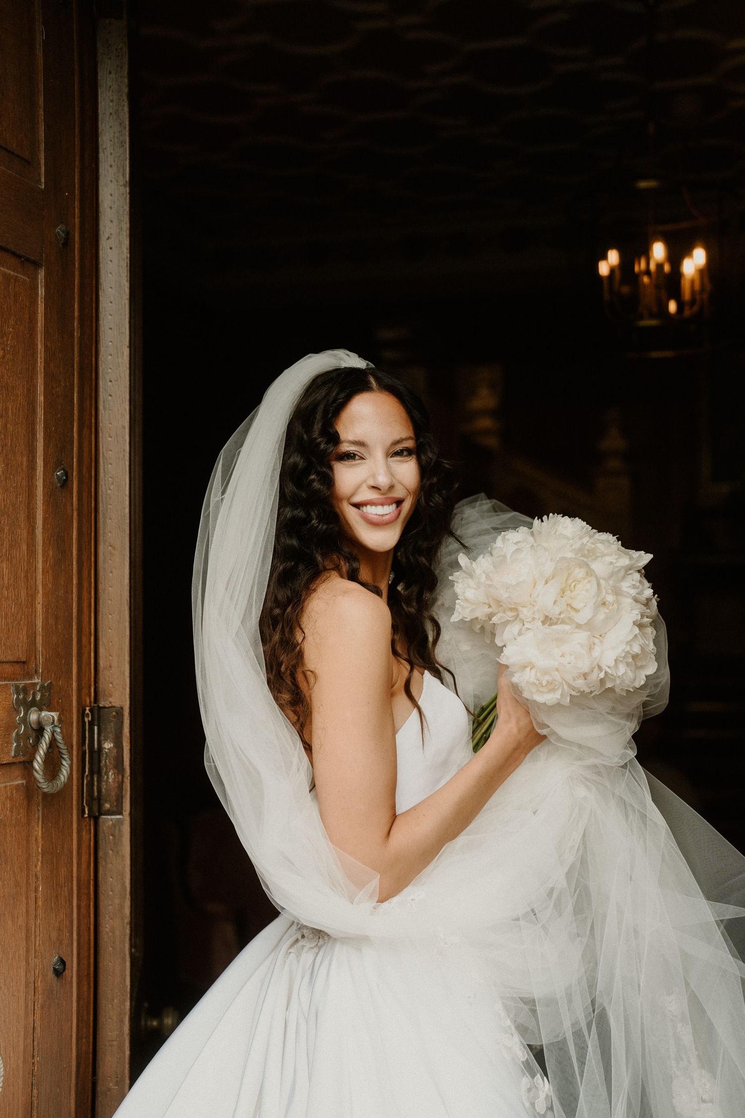 Bride in flowing white gown holds bouquet while smiling in warm indoor lighting.