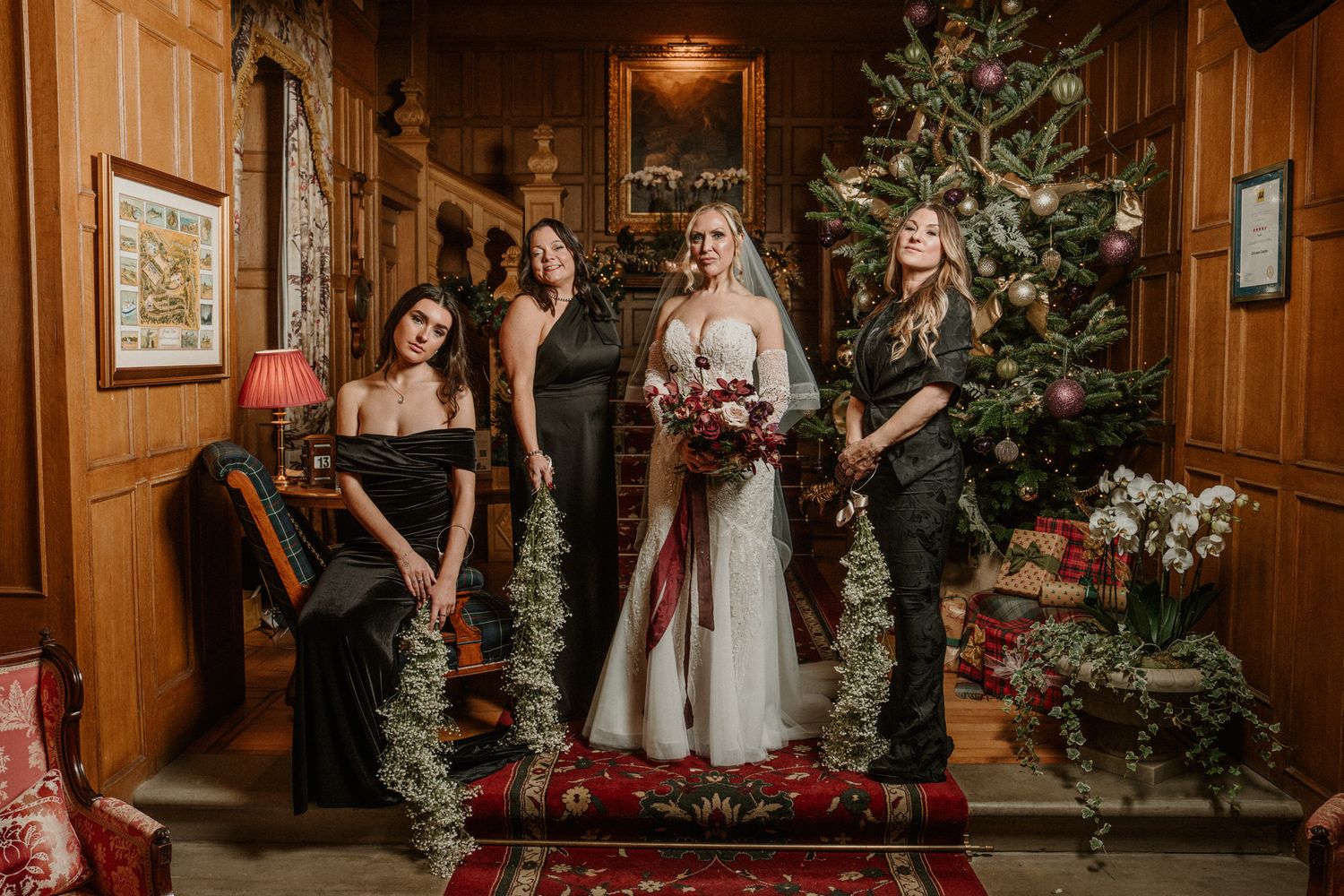 Bridal party stands on red-carpeted stairs in wood-paneled room with Christmas decorations and evergreen garlands.