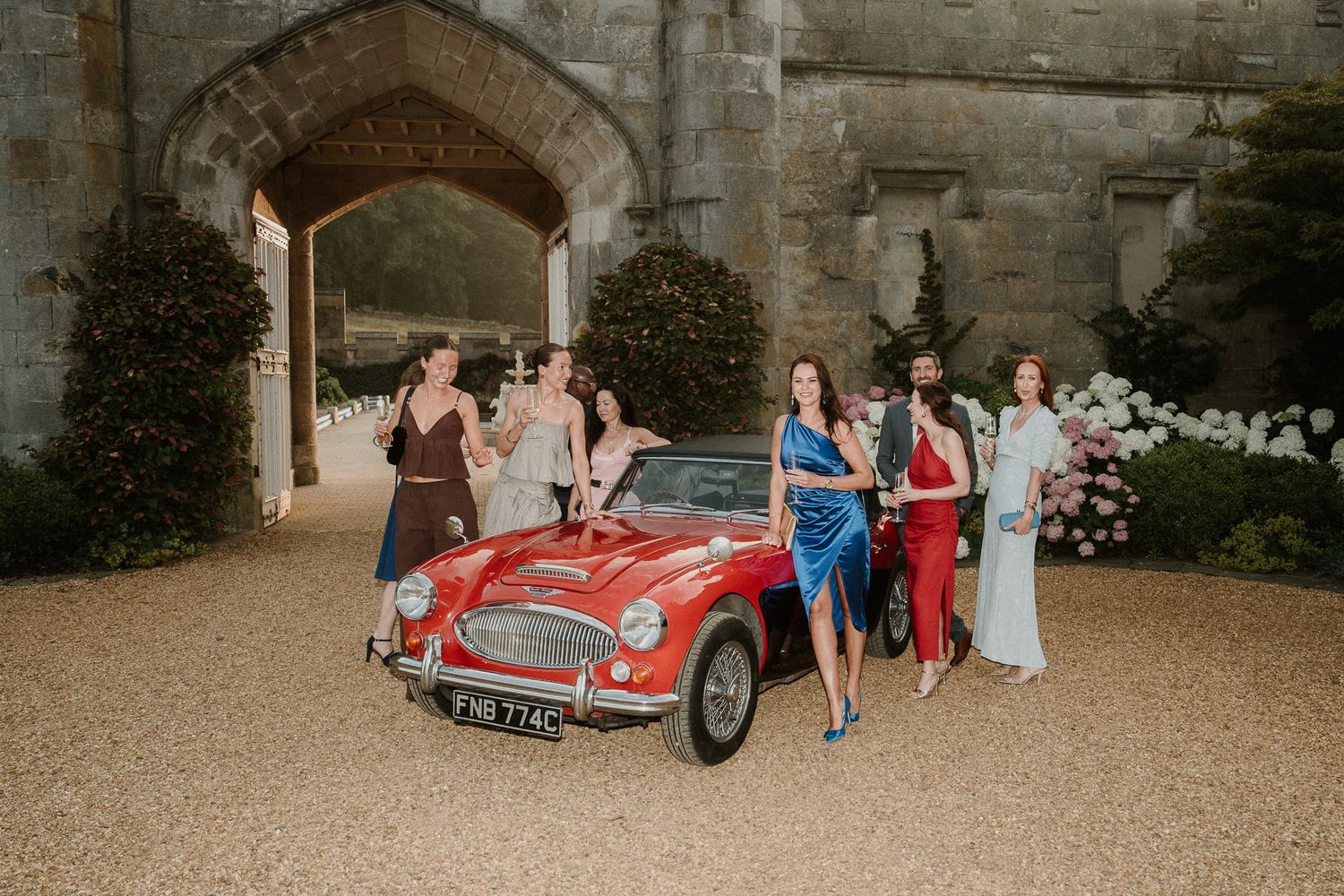 A classic red vintage car is displayed with a wedding party under a stone archway at an elegant venue.