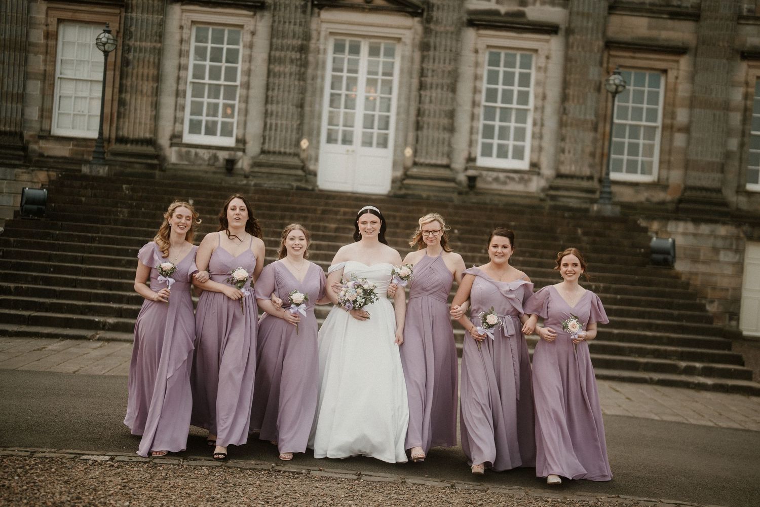 Wedding party in lavender bridesmaid dresses poses on grand stone steps of historic building.
