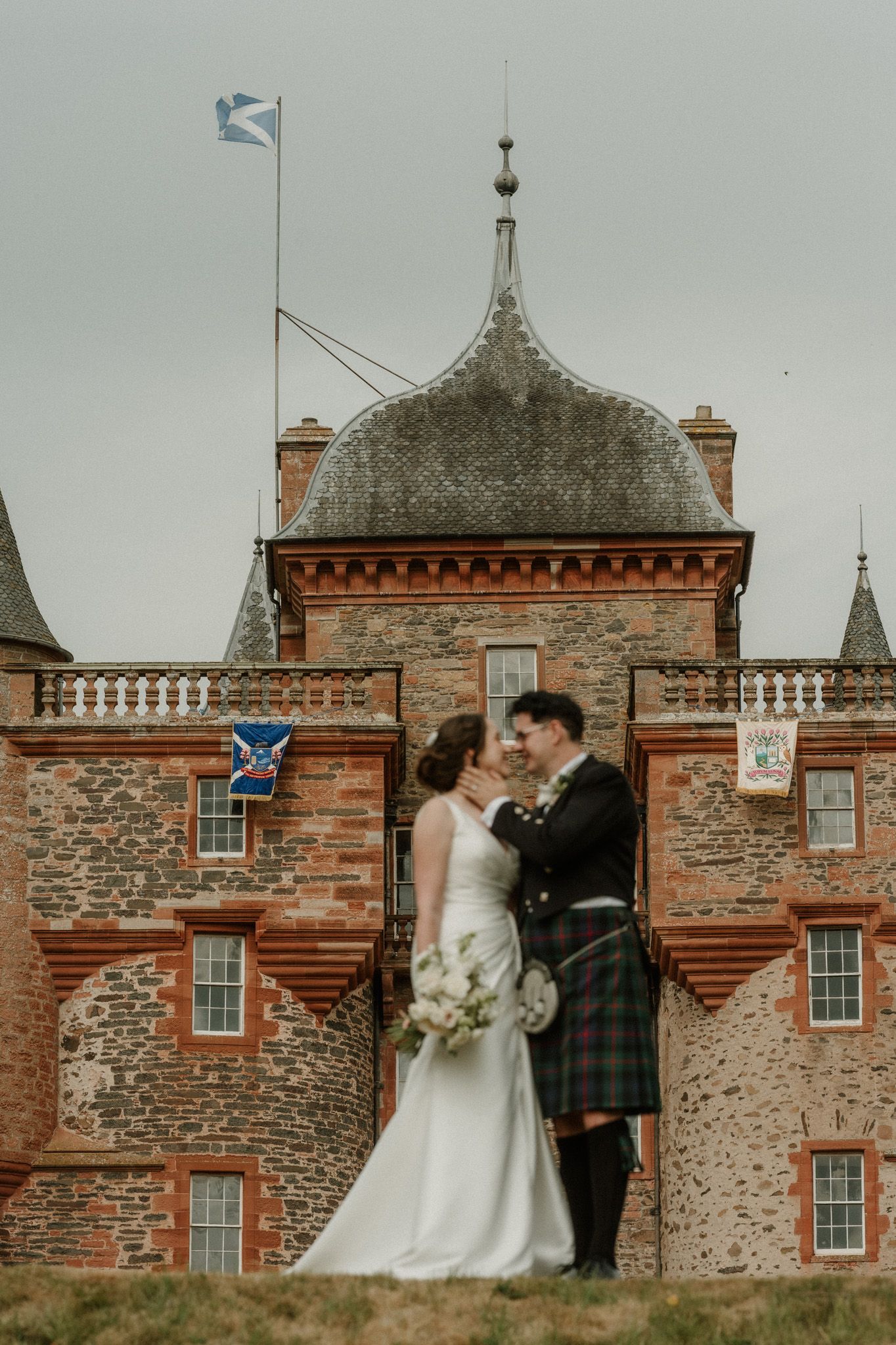 Bride in a white gown and groom in a tartan kilt share an intimate moment before a historic red sandstone Scottish castle with a flag.