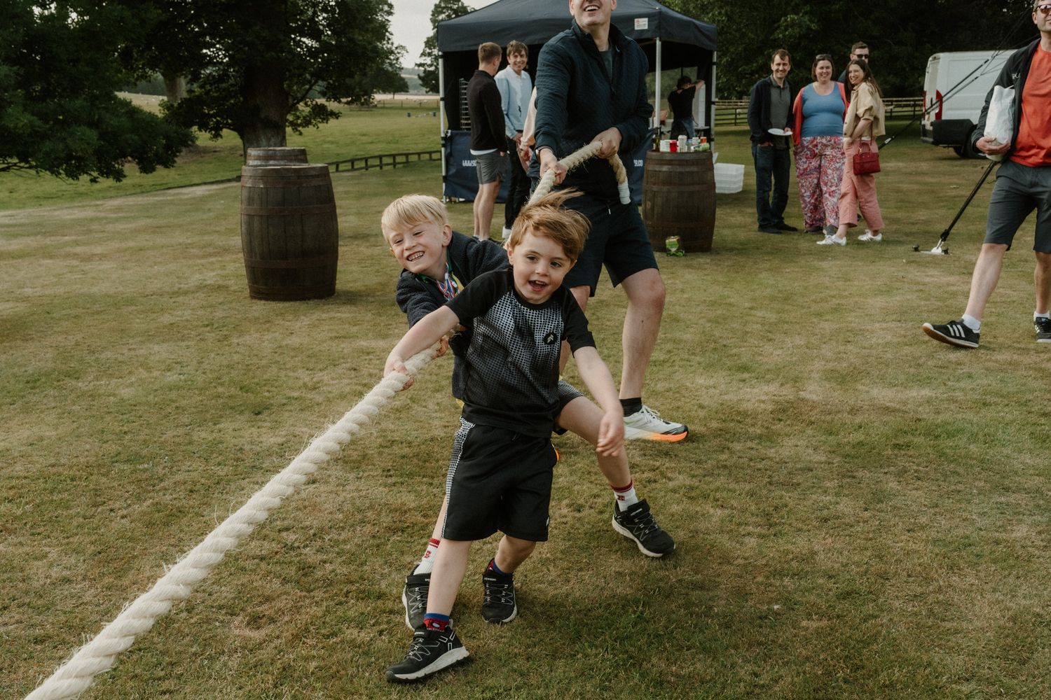 Two young boys in dark clothes pull hard on a thick rope during a tug of war game on a grassy lawn at an outdoor event.