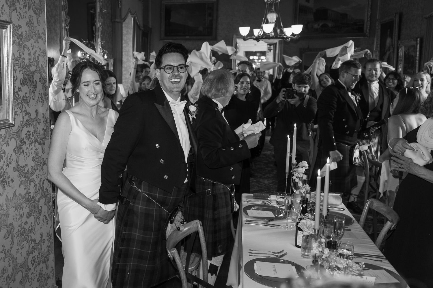 Bride in white gown and groom in tartan kilt walk through a wedding reception as guests wave napkins beside a candlelit table.