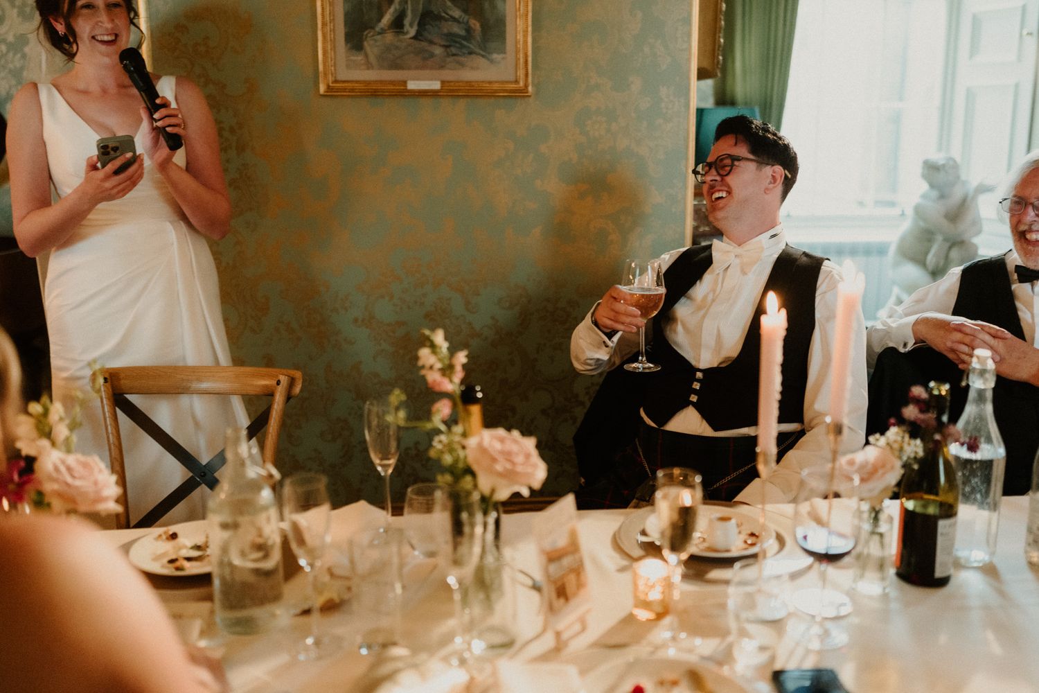 Woman in a white dress holds a microphone and gives a toast while seated wedding guests raise glasses at a candlelit reception table.