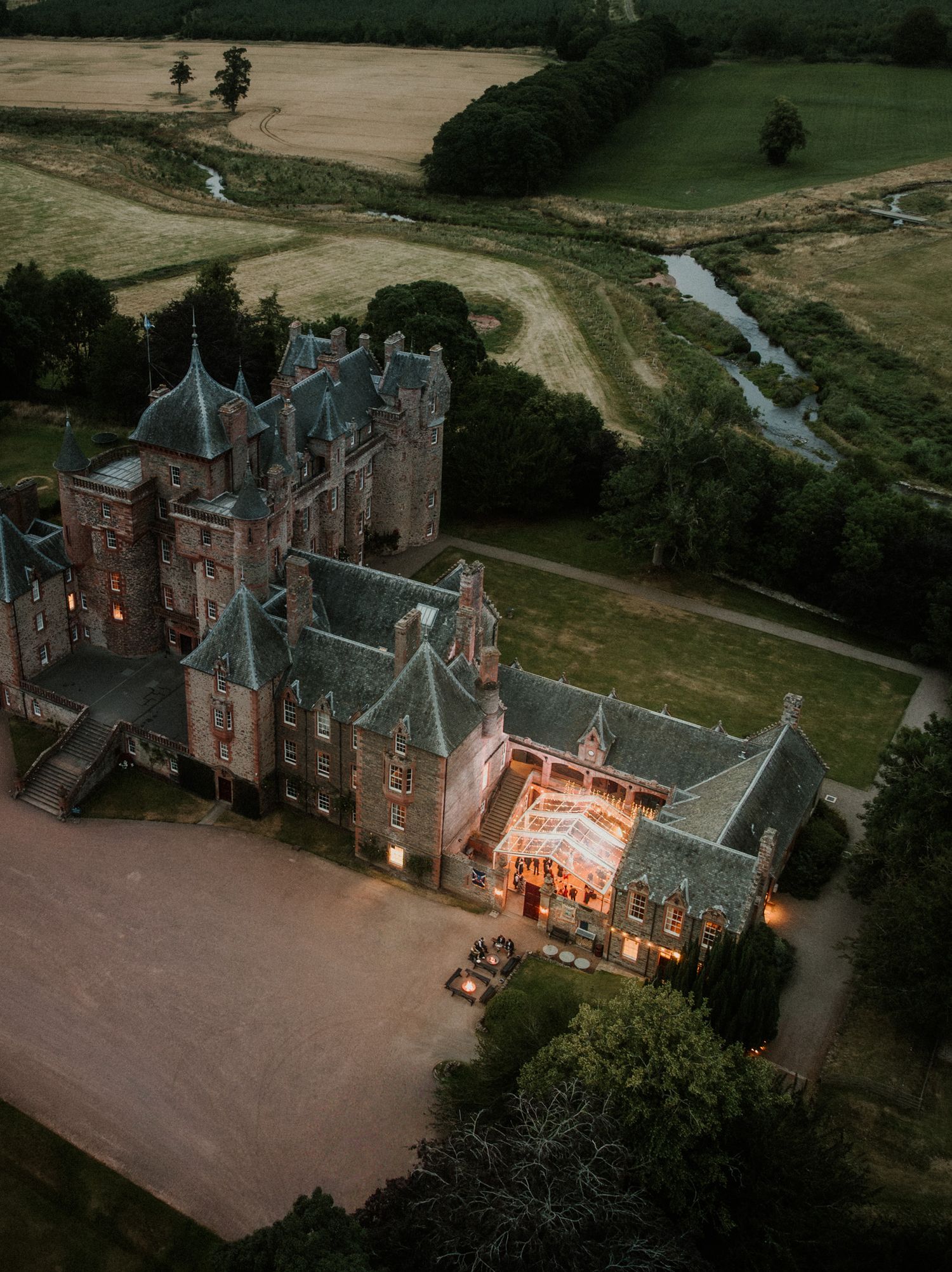 Aerial view at dusk of a grand turreted stone castle with a glowing illuminated pavilion, surrounded by lawns, trees and a winding stream.
