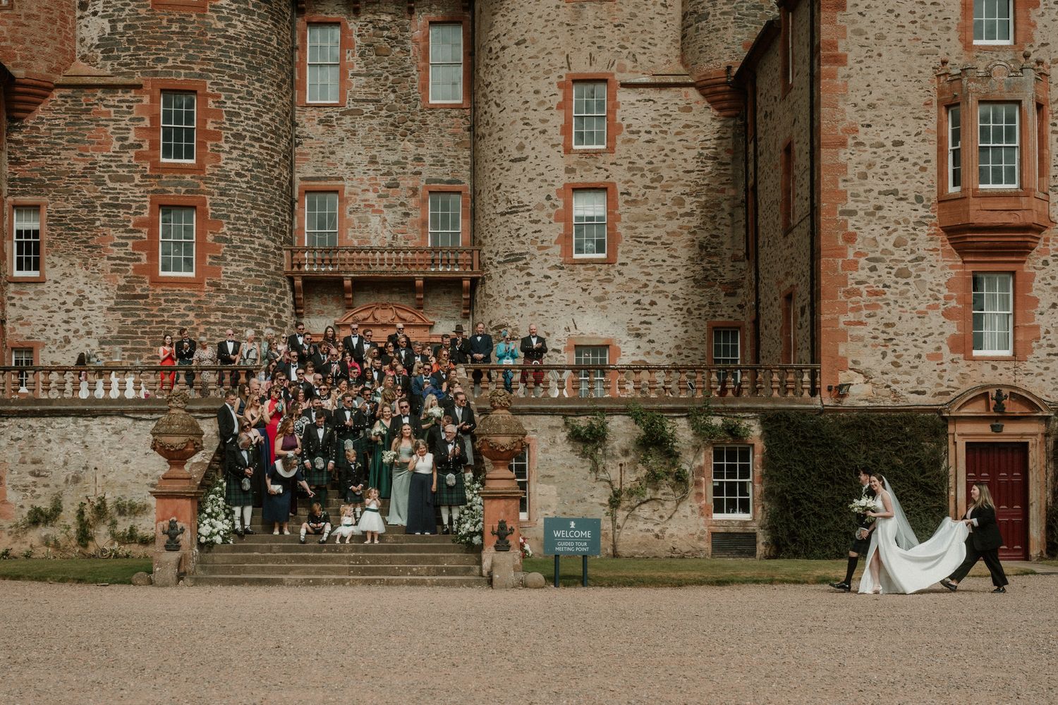 Wedding guests gather on the stone steps of a historic castle while a bride in a white gown and groom walk across the gravel courtyard.
