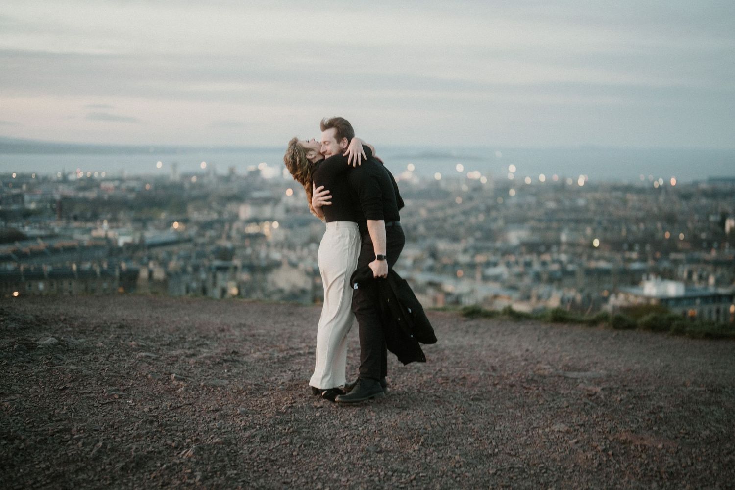 A couple sharing a passionate kiss on a hilltop at dusk with a glowing city skyline behind them.