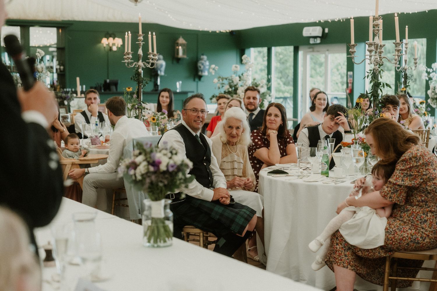 Wedding guests smile and celebrate at reception tables decorated with purple floral centerpieces in green venue.
