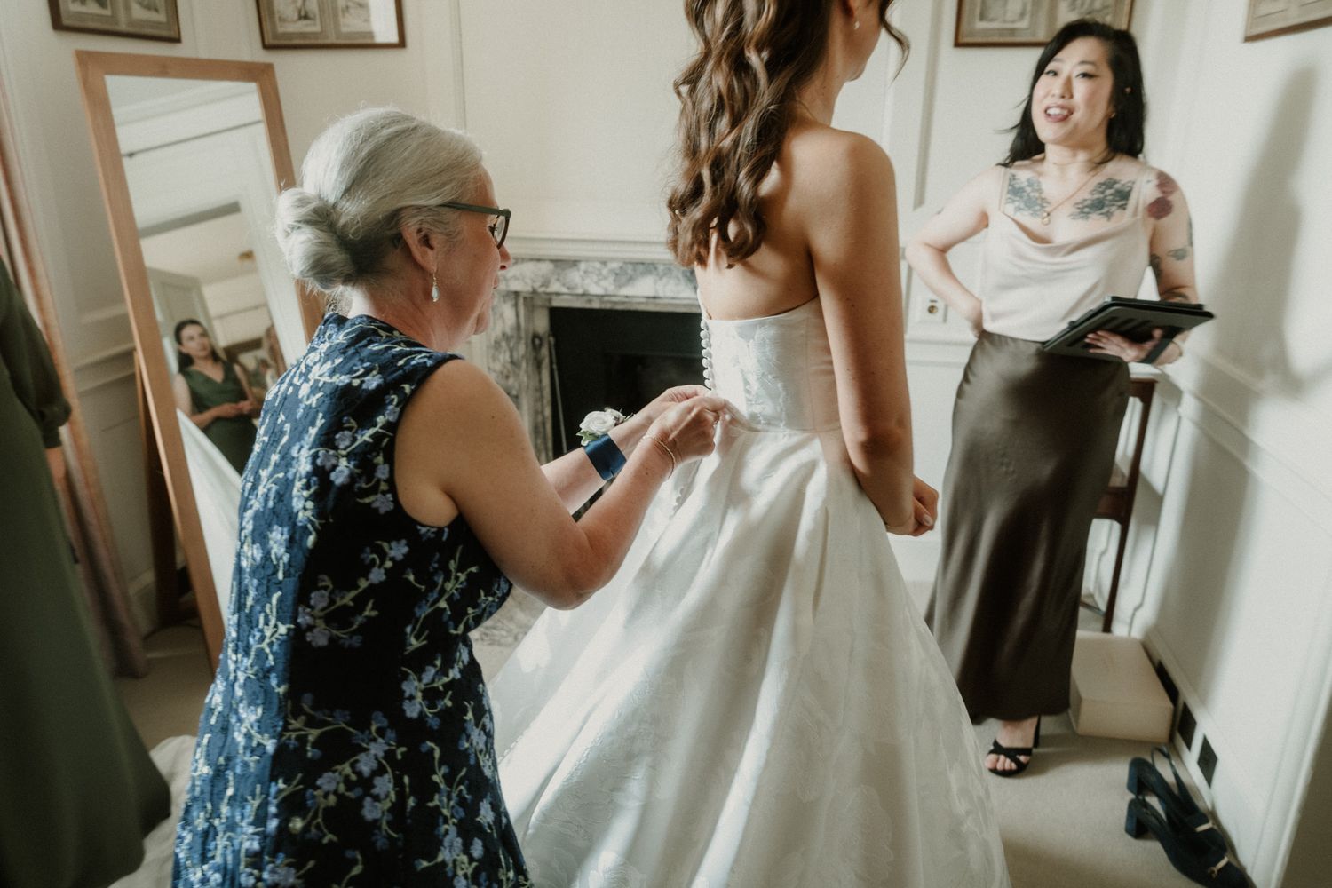 A bride in a white gown being helped by an older woman while a tattooed guest watches in an elegant room.